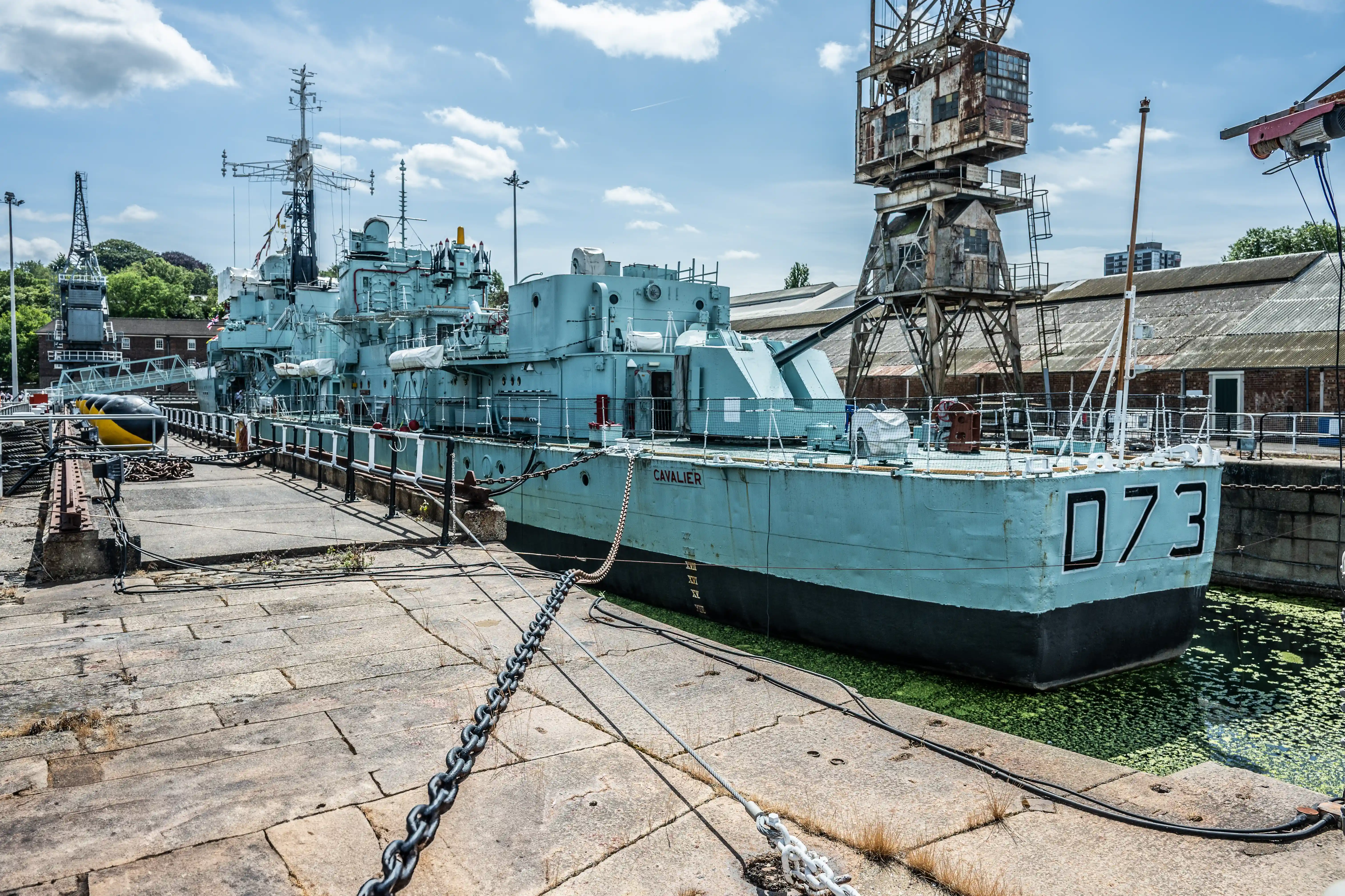 The image depicts a docked naval ship, specifically a destroyer, with the number 073 painted on its bow. The ship is moored to a concrete pier with chains and ropes, and there is a rusty, industrial-looking crane structure in the background. The ship appears to be part of a historical or decommissioned fleet, given the setting and the presence of other similar vessels in the vicinity. The overall scene suggests a maritime or naval museum setting.