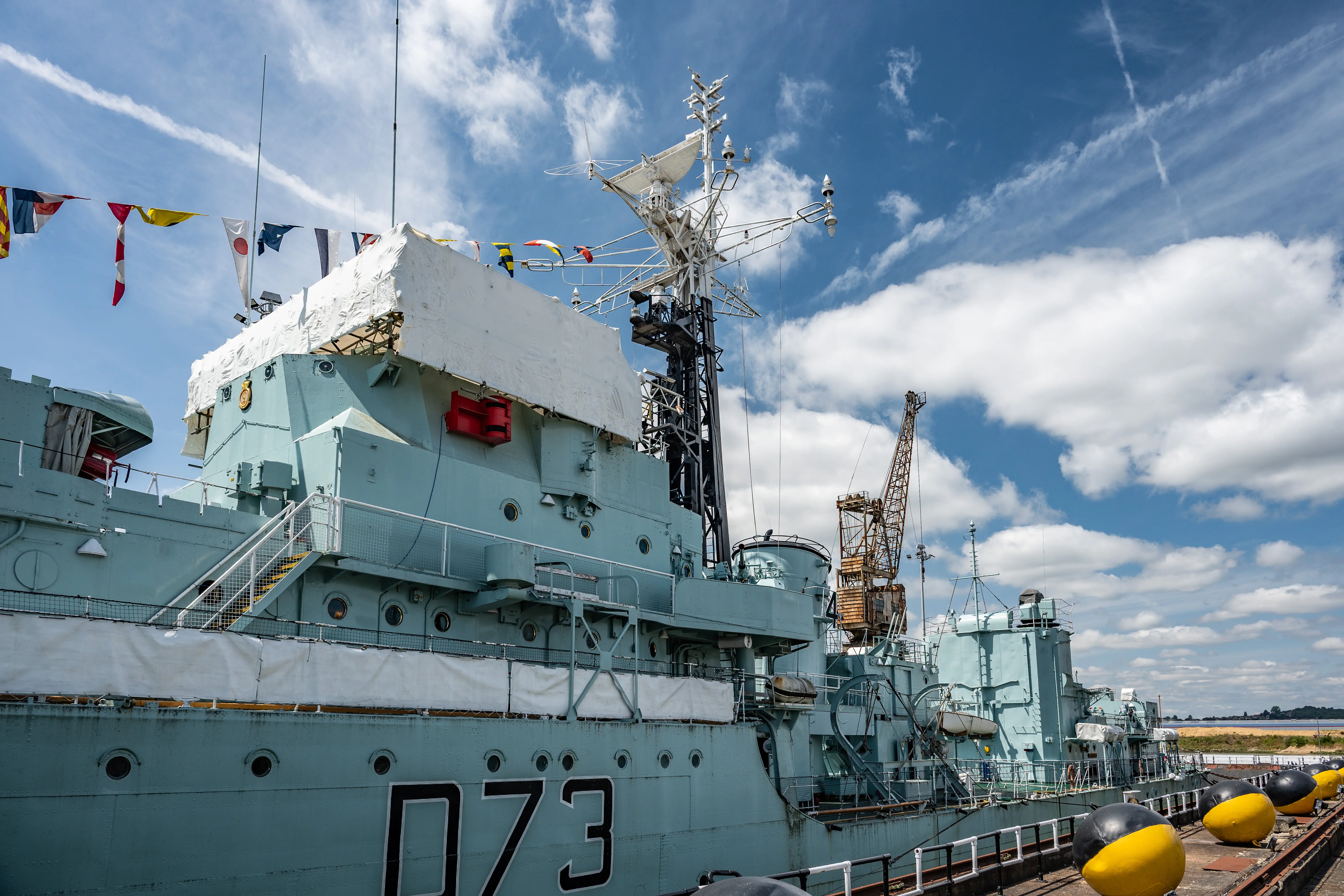 The image depicts a large naval ship docked at a port. The ship is painted in a light blue-gray color and has the identification number '073' prominently displayed on its side. Various flags are flying from the ship, and it is equipped with multiple antennas and radar equipment. The ship appears to be well-maintained and is situated under a partly cloudy sky.