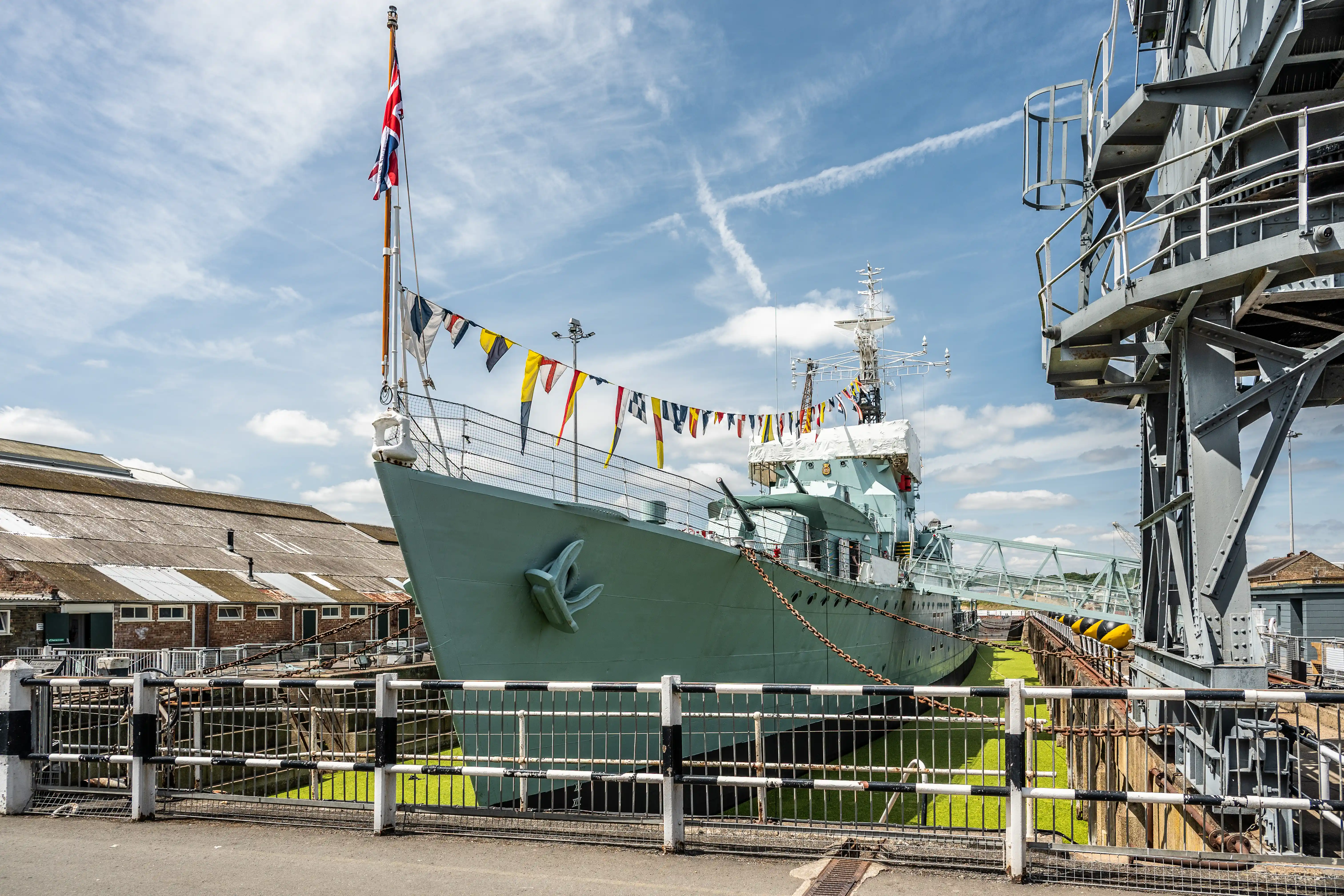 The image depicts a large naval ship docked at a port. The ship is adorned with various signal flags and a prominent Union Jack flag at the stern. The ship's hull is painted in a light green color, and it features an anchor emblem on the bow. The dock area includes railings, a staircase, and industrial buildings in the background under a partly cloudy sky.