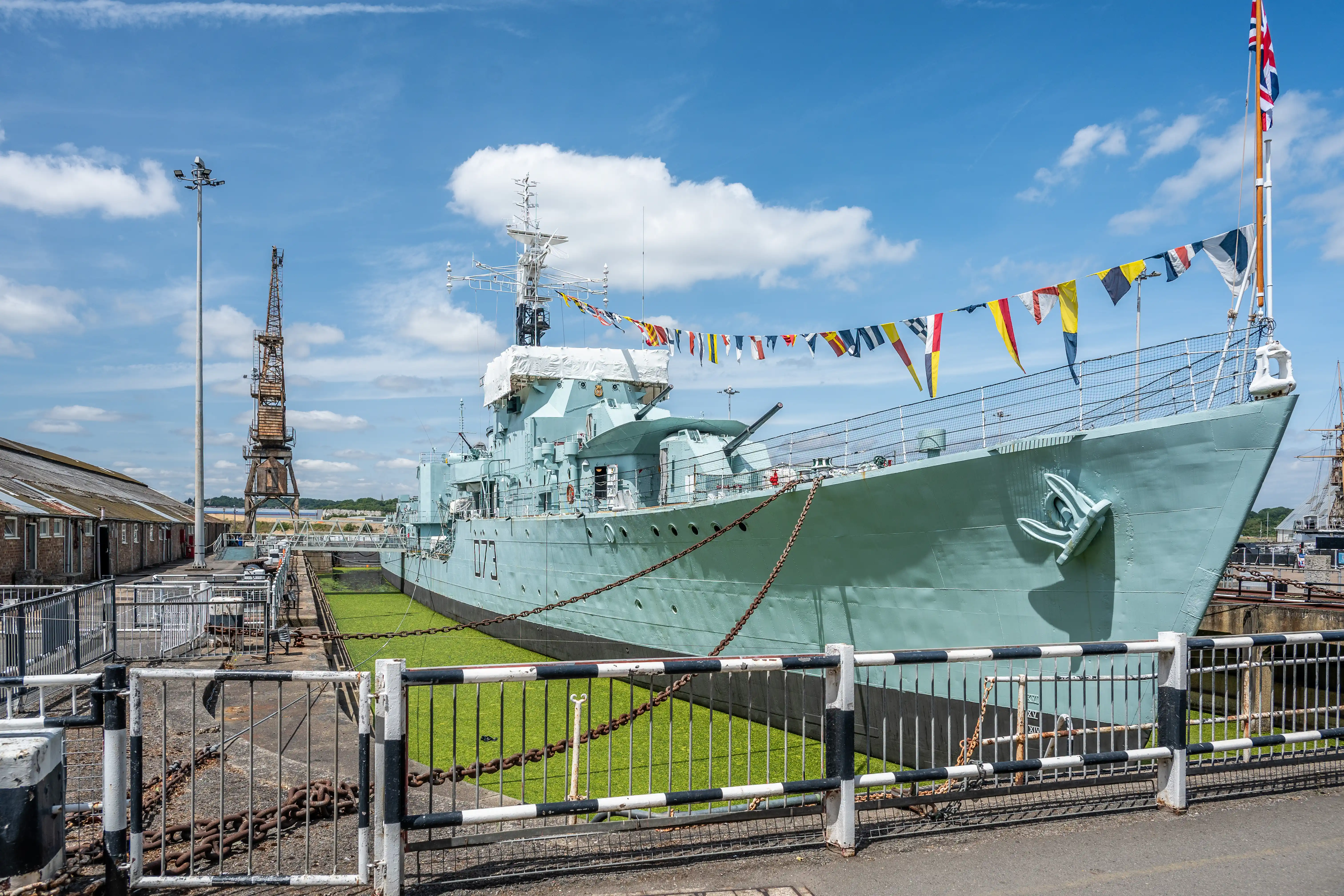 The image depicts a large naval ship docked at a harbor. The ship is adorned with numerous colorful signal flags and has a prominent anchor visible on its side. The surrounding area includes a fenced walkway, a crane structure, and industrial buildings. The sky is clear with a few scattered clouds, indicating a sunny day.