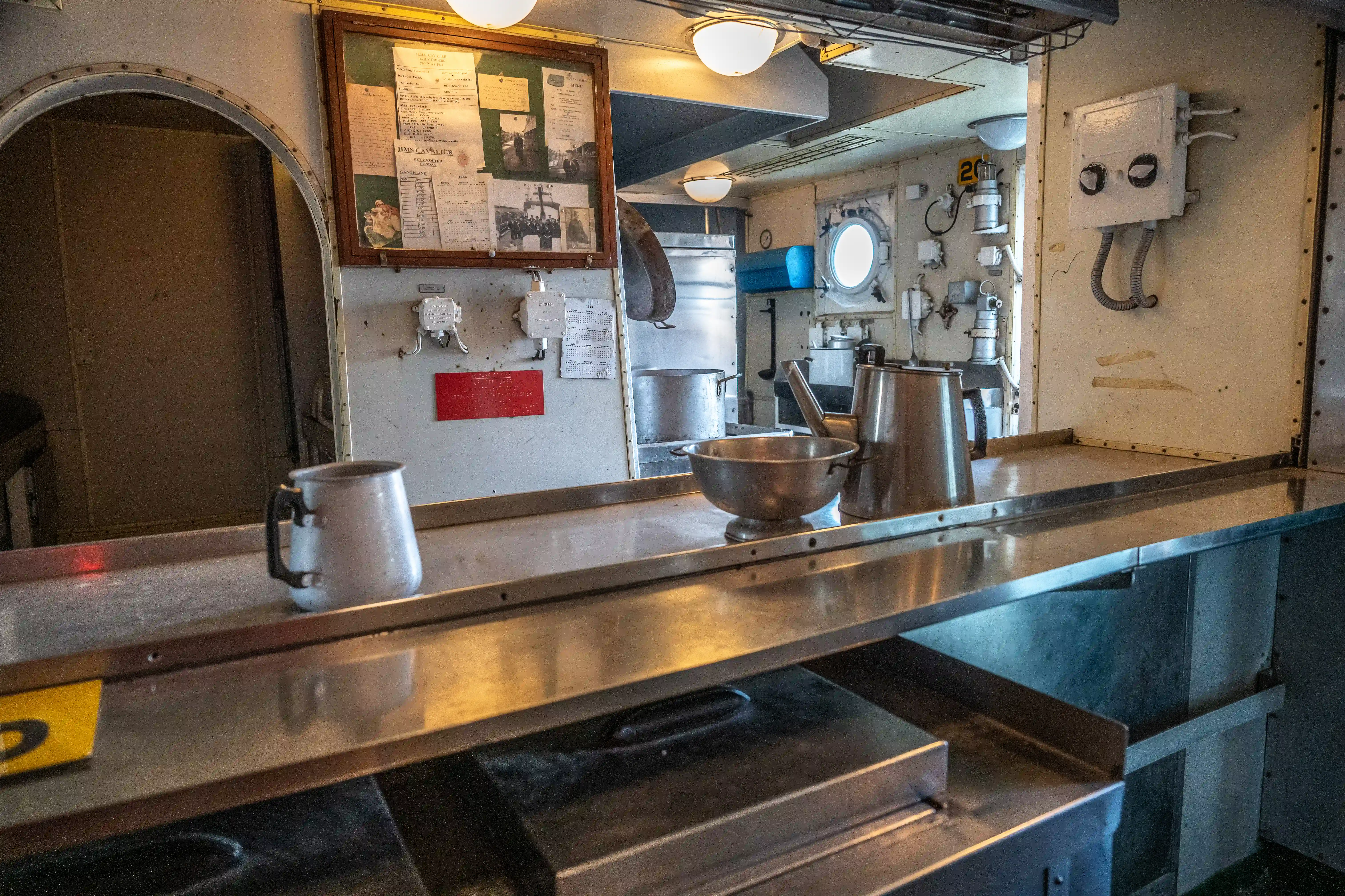 The image depicts the interior of a ship's galley or kitchen. The space is compact and utilitarian, featuring stainless steel countertops, a large metal bowl, a kettle, and various kitchen utensils. There is a bulletin board with several documents and photographs pinned to it, and a round porthole that allows natural light to enter. The walls and equipment suggest a functional and efficient workspace designed for preparing meals at sea.