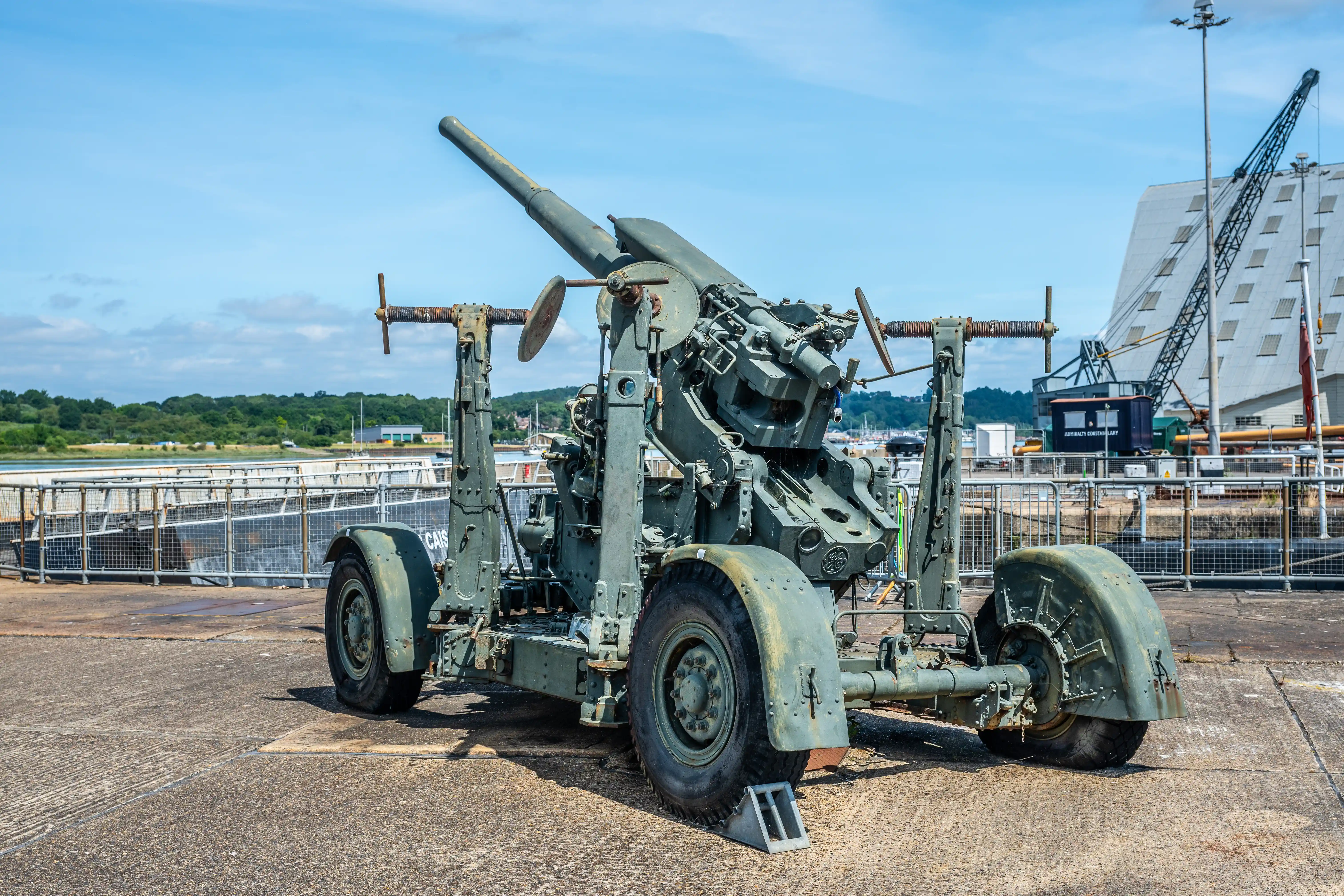 The image depicts a large, mounted artillery gun situated on a concrete surface near a dock or port area. The gun is elevated on a wheeled carriage and appears to be of historical military significance. In the background, there is a modern building with a unique architectural design, cranes, and a scenic view of greenery and water, suggesting the location might be a historical site or museum near a harbor.