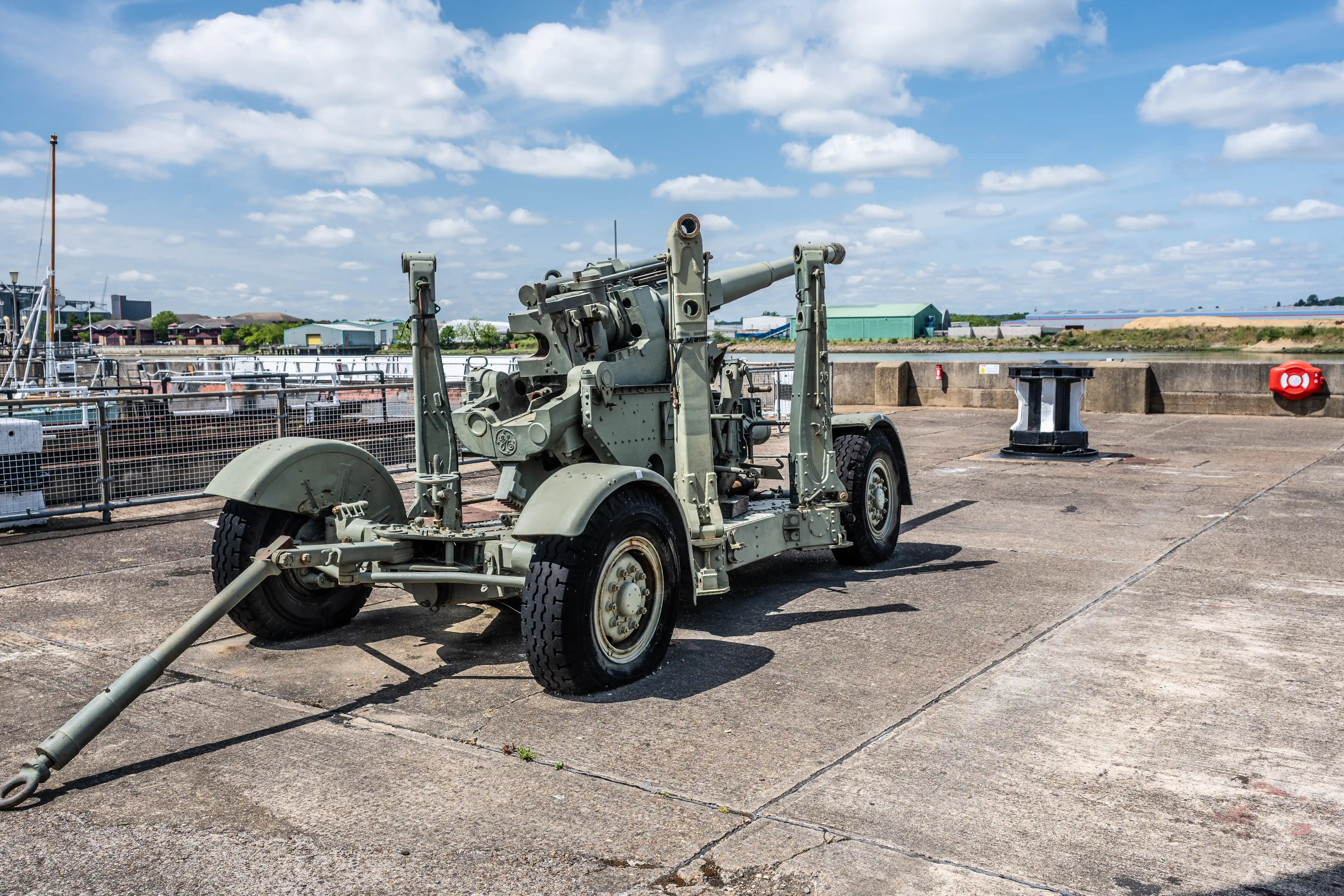 The image depicts a military artillery piece positioned on a concrete surface near a waterfront. The artillery is mounted on a mobile carriage with large wheels and a towing hitch, suggesting it is designed for transport. The background includes a body of water, industrial buildings, and a partly cloudy sky. The setting appears to be a dock or a port area.