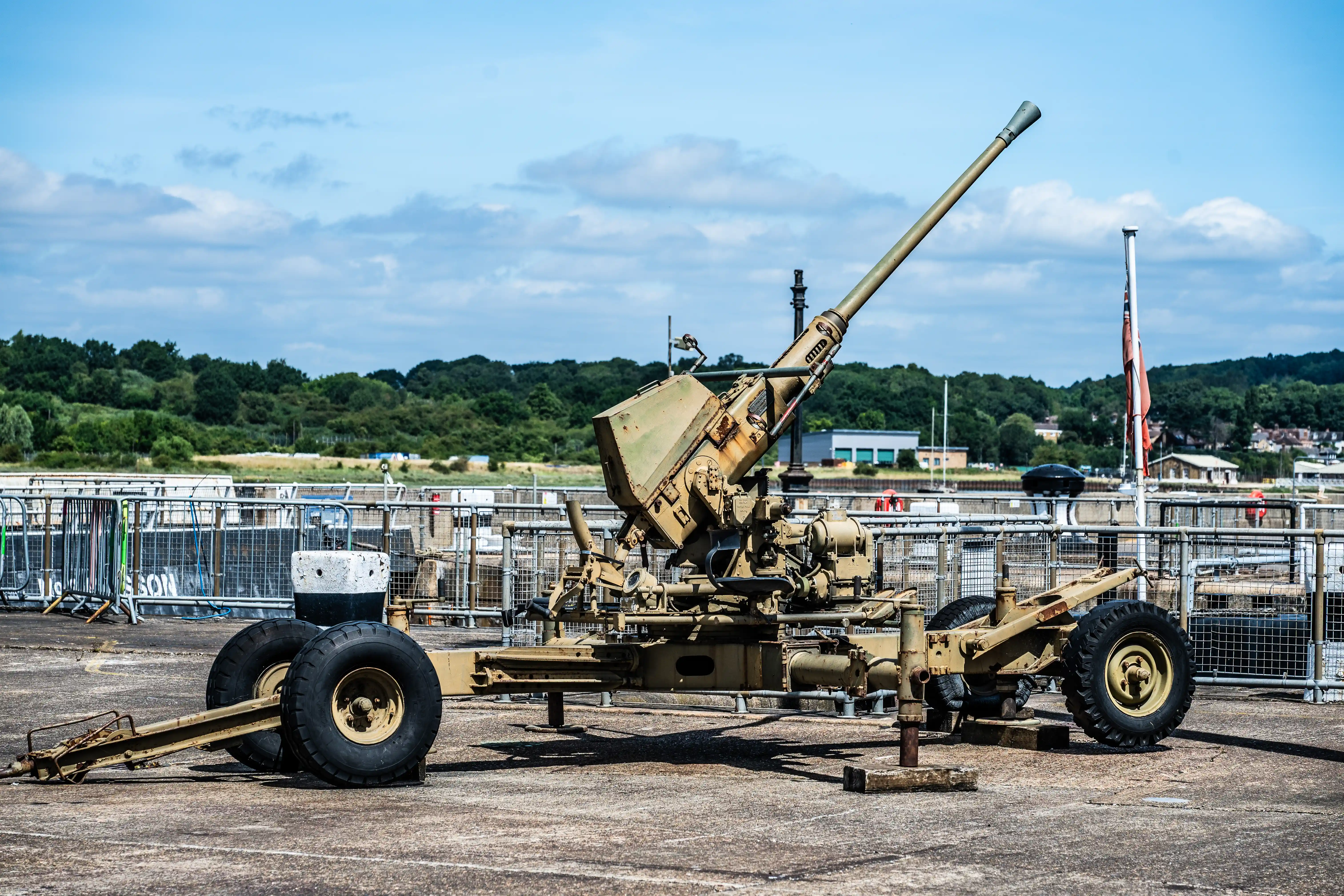 The image depicts a large military artillery piece, specifically an anti-aircraft gun, mounted on a wheeled carriage. It is situated on a paved area, likely a dock or a similar open space, with a backdrop of greenery and distant buildings. The gun is positioned on a platform with barriers and fencing around it, suggesting it might be part of a historical display or museum exhibit. The overall scene is set under a clear, blue sky with scattered clouds.