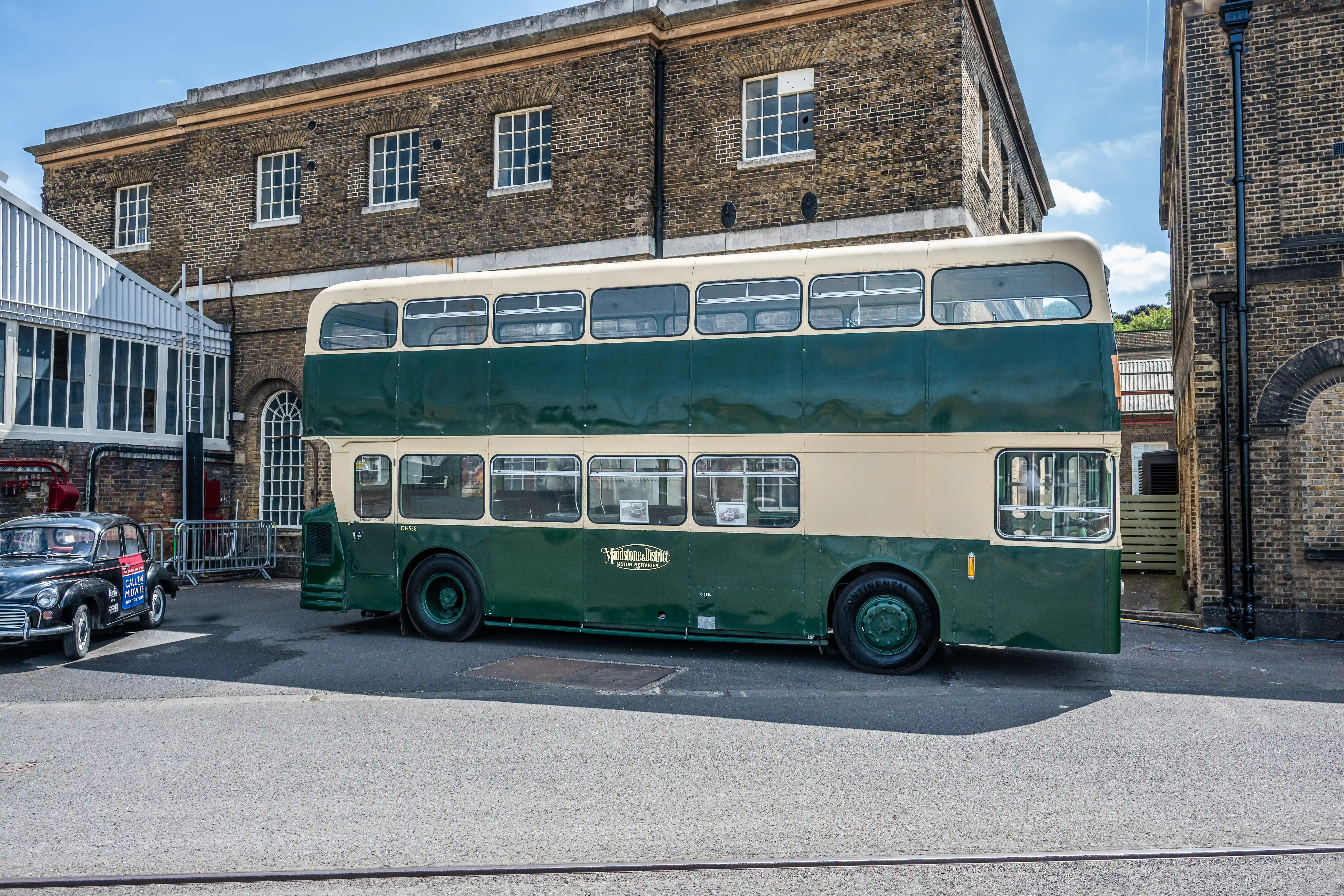 The image depicts a vintage double-decker bus painted in green and cream colors, parked in an urban setting. The bus is situated in front of a brick building with multiple windows, and there is another vintage car parked nearby. The scene appears to be part of a historical or museum exhibit, showcasing classic vehicles.