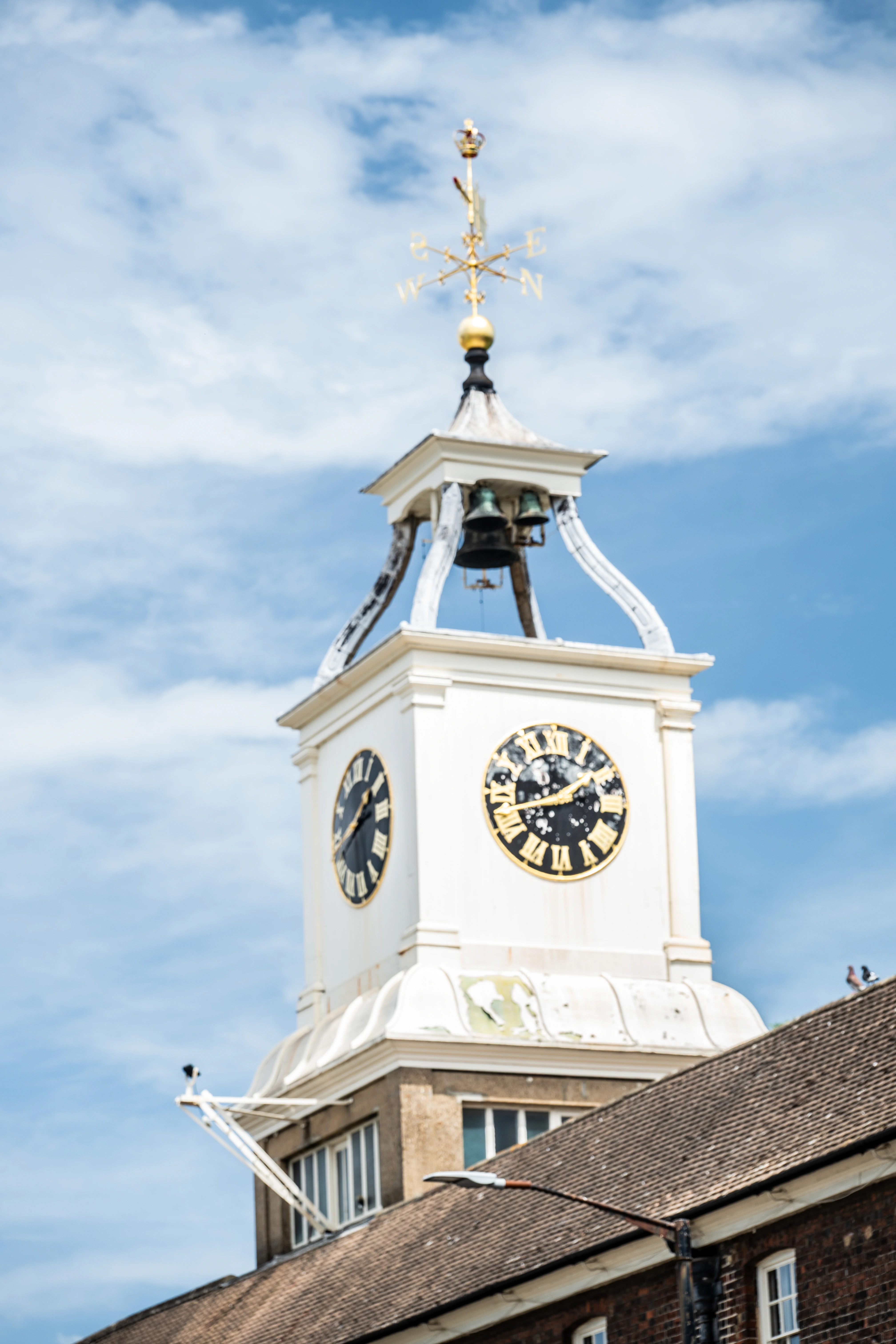 The image depicts a clock tower with a weather vane on top, showing the directions. The tower has two visible clock faces with Roman numerals. The structure is white and appears to be part of a larger building with a tiled roof. The sky in the background is partly cloudy.