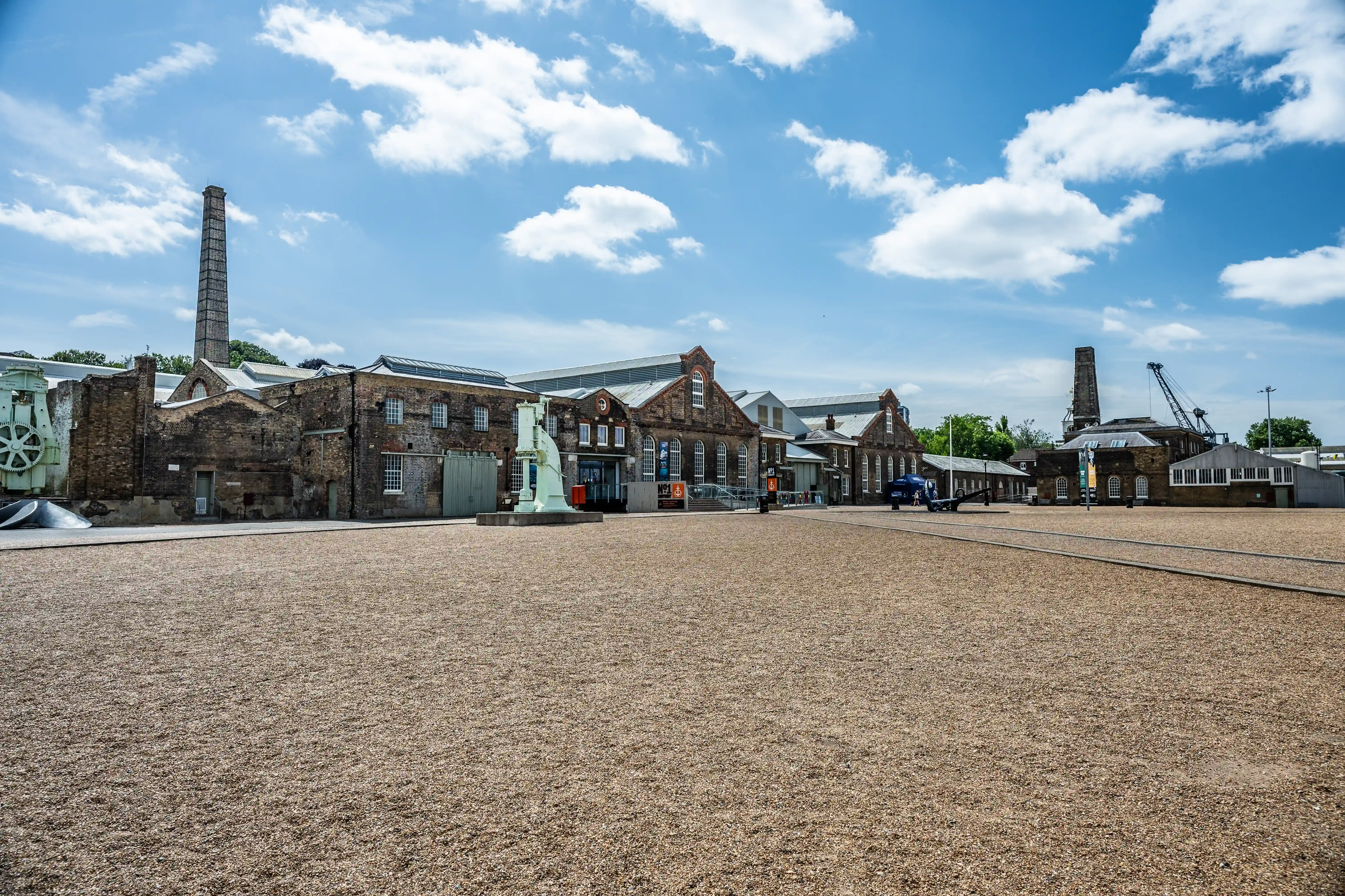 The image depicts an industrial site with historical brick buildings, a large open gravel area, and remnants of old industrial equipment such as a tall chimney and machinery. The sky is clear with scattered clouds, suggesting a pleasant weather. The buildings have a mix of old and slightly modernized architecture, indicating a preserved historical site, possibly a museum or heritage site. The overall scene conveys a sense of historical significance and industrial heritage.
