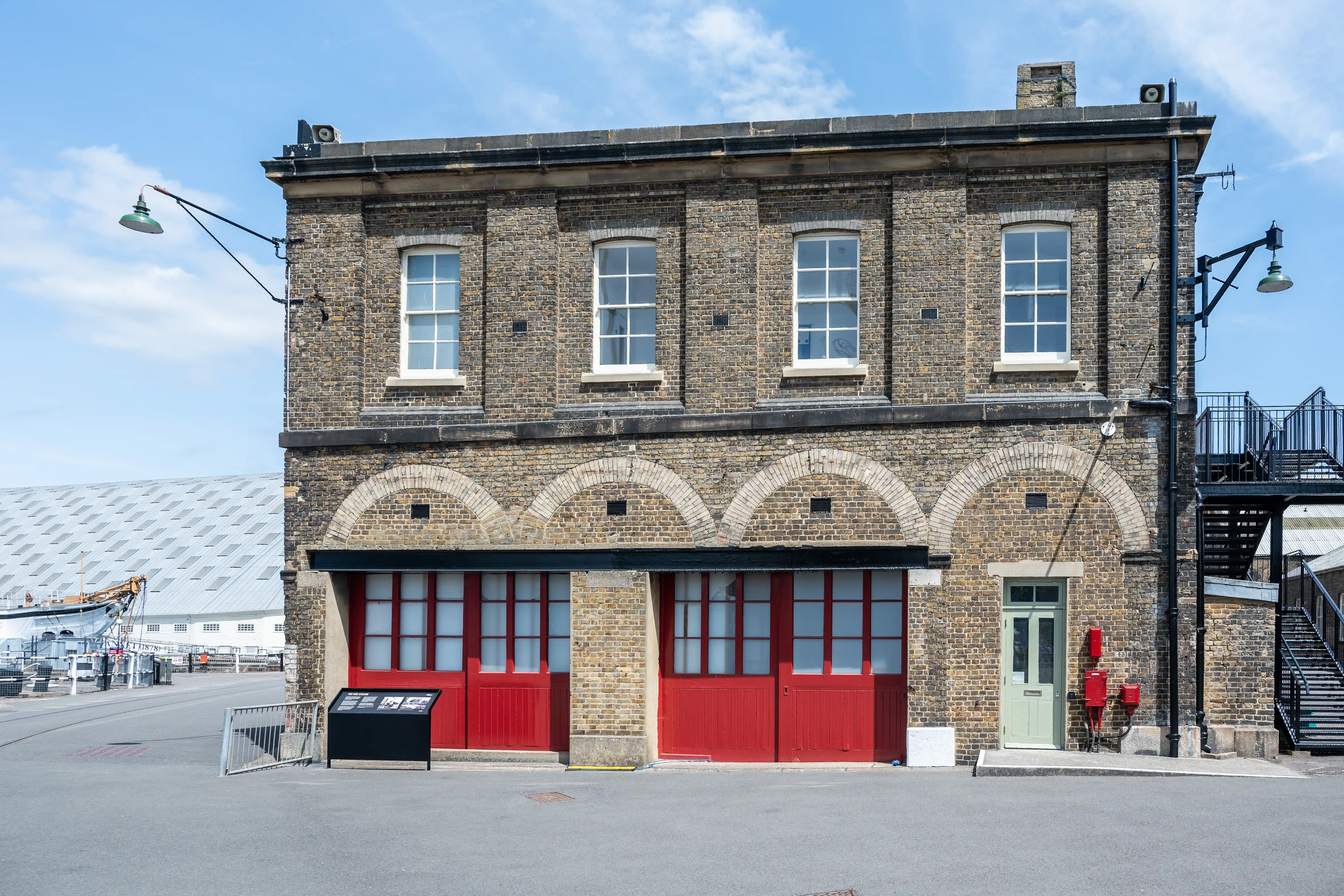 The image depicts a two-story brick building with a distinct architectural style. The building features large, red double doors on the ground floor and several evenly spaced windows on the upper floor. There are arched brick details above the ground floor and a metal staircase on the right side. The building is situated in an open area with a clear sky and some industrial elements visible in the background.