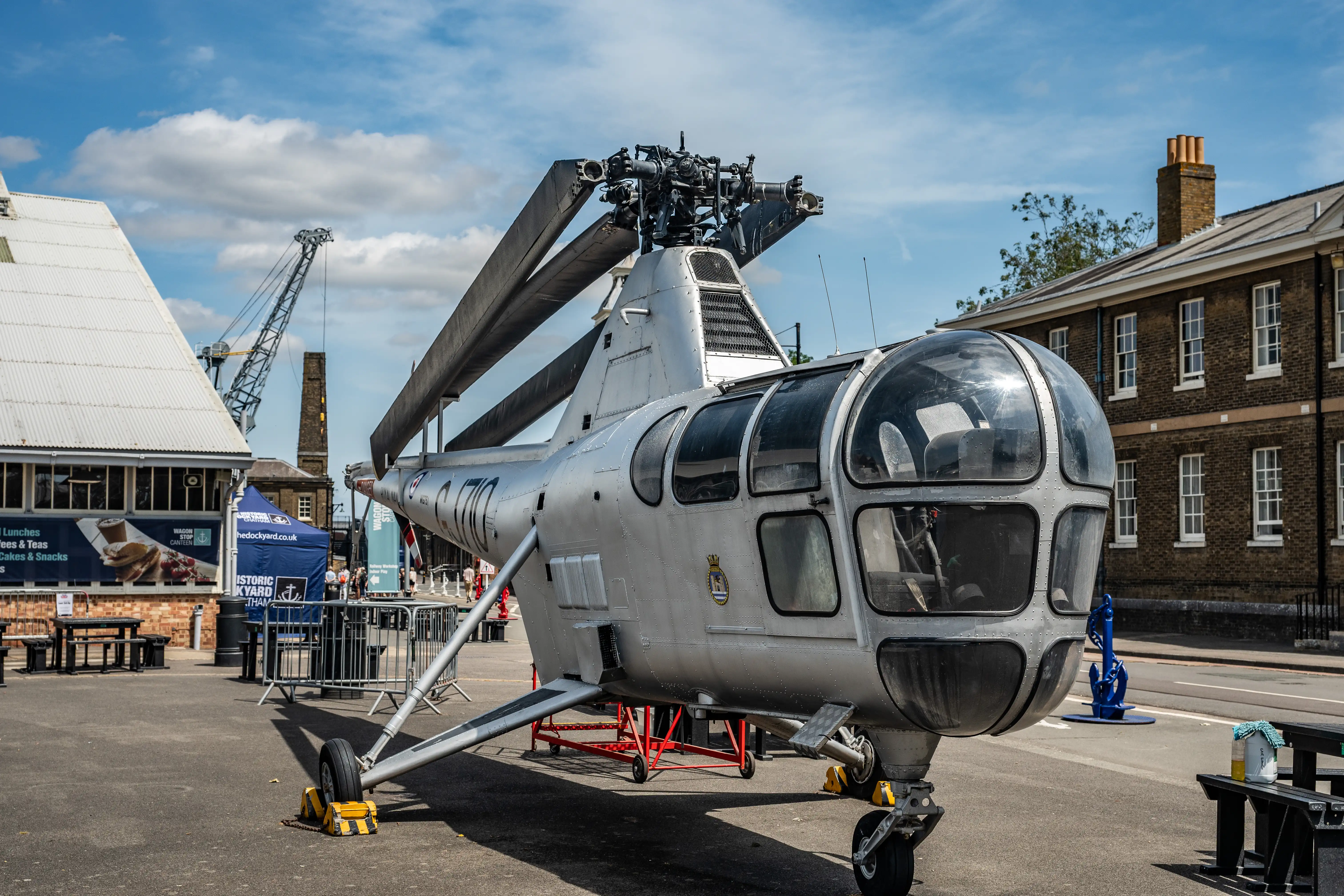 The image depicts a vintage helicopter on display at an outdoor event. The helicopter is positioned in an open area with buildings and a crane in the background. There are informational signs and barriers around the display area, indicating it is part of an exhibition or museum. The weather is clear with a blue sky and some clouds. The setting suggests a historical or educational context, likely related to aviation or military history.