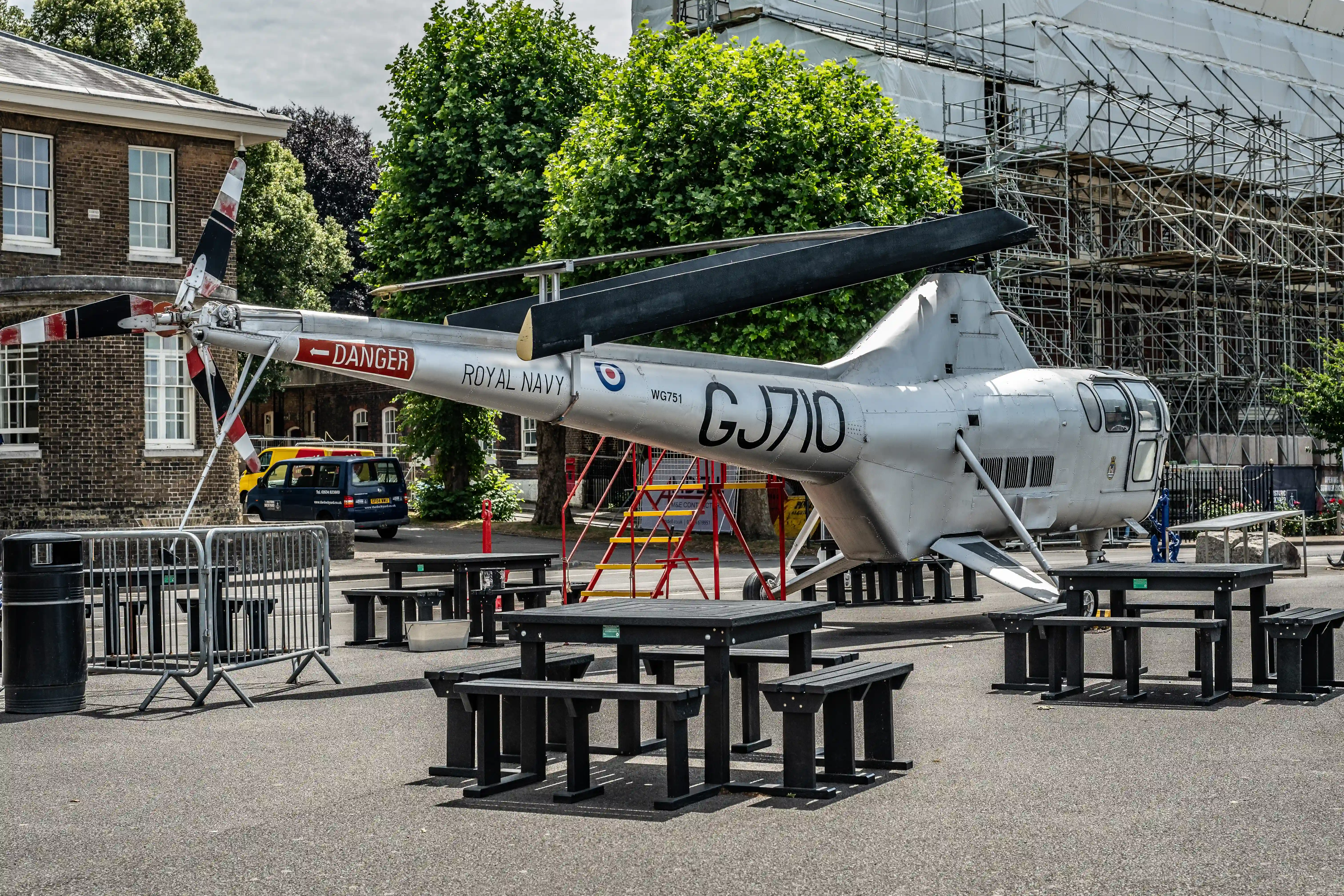 The image depicts a Royal Navy helicopter on display in an outdoor setting. The helicopter is marked with the identification 'GJ710' and has a 'DANGER' sign on its tail rotor. It is surrounded by several black picnic tables and benches, with barriers and scaffolding visible in the background. The scene appears to be part of an exhibition or museum display.