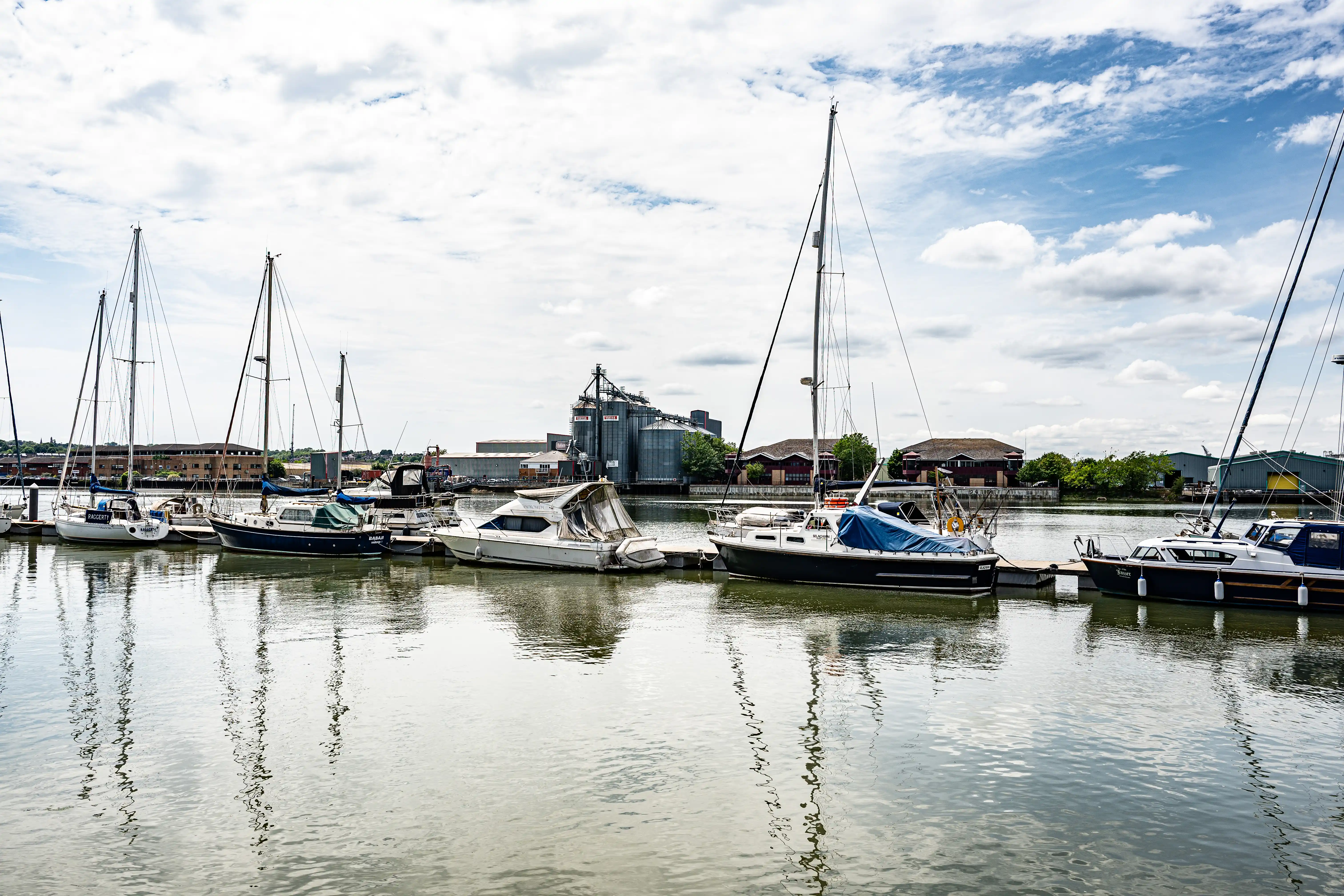 The image depicts a serene marina with several sailboats and yachts docked in calm water. The boats are moored in an orderly fashion, and their reflections are visible in the water. In the background, there are industrial buildings, including large silos and warehouses, suggesting a nearby port or industrial area. The sky is partly cloudy with patches of blue, adding to the tranquil atmosphere of the scene.