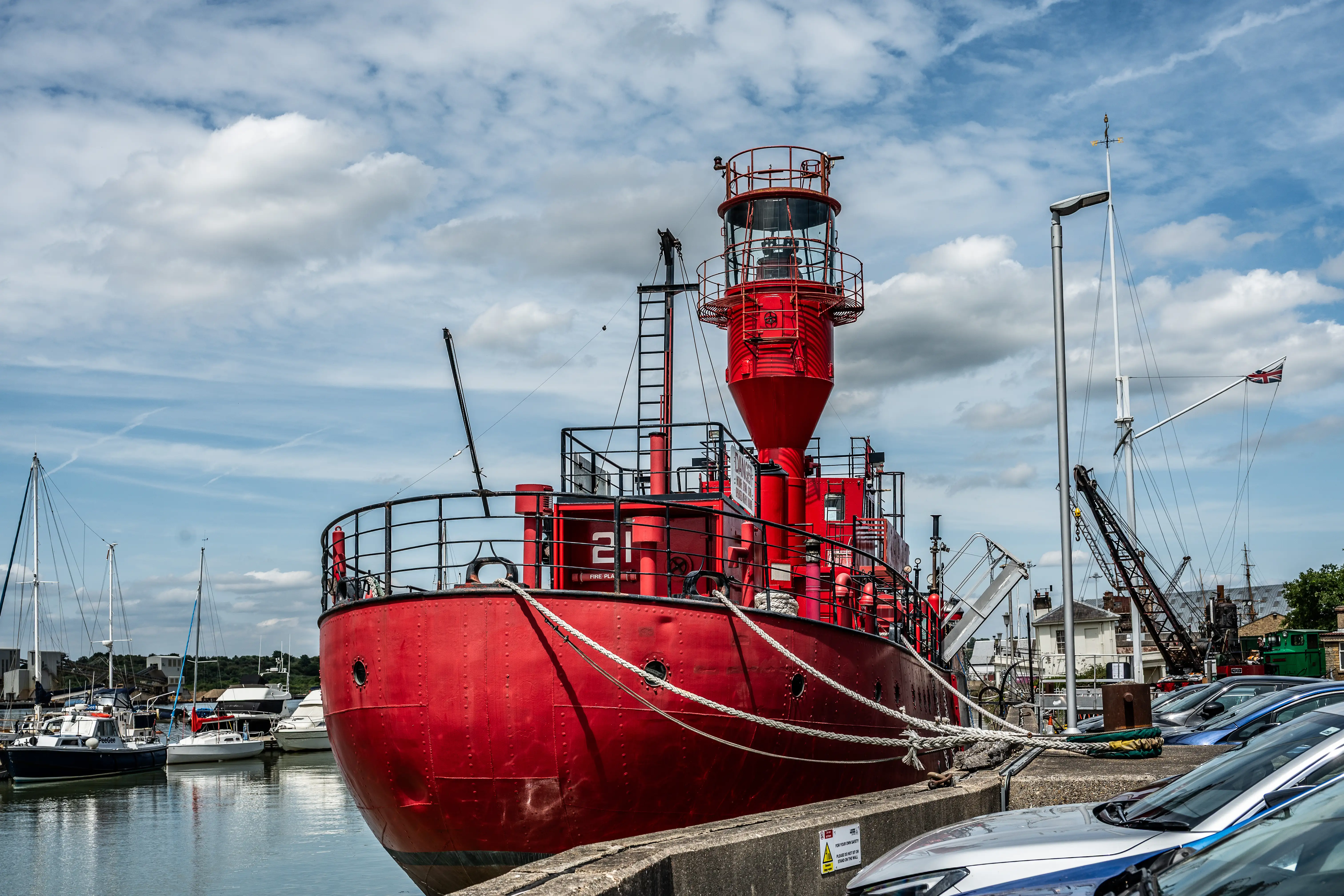 The image depicts a vibrant red ship docked at a marina. The ship is prominently marked with the number 21 and has a distinctive light tower structure on its deck. Surrounding the ship are other boats and yachts, as well as a parking area with several cars. The sky is partly cloudy, and a flag is visible in the background, indicating a likely coastal or harbor setting.