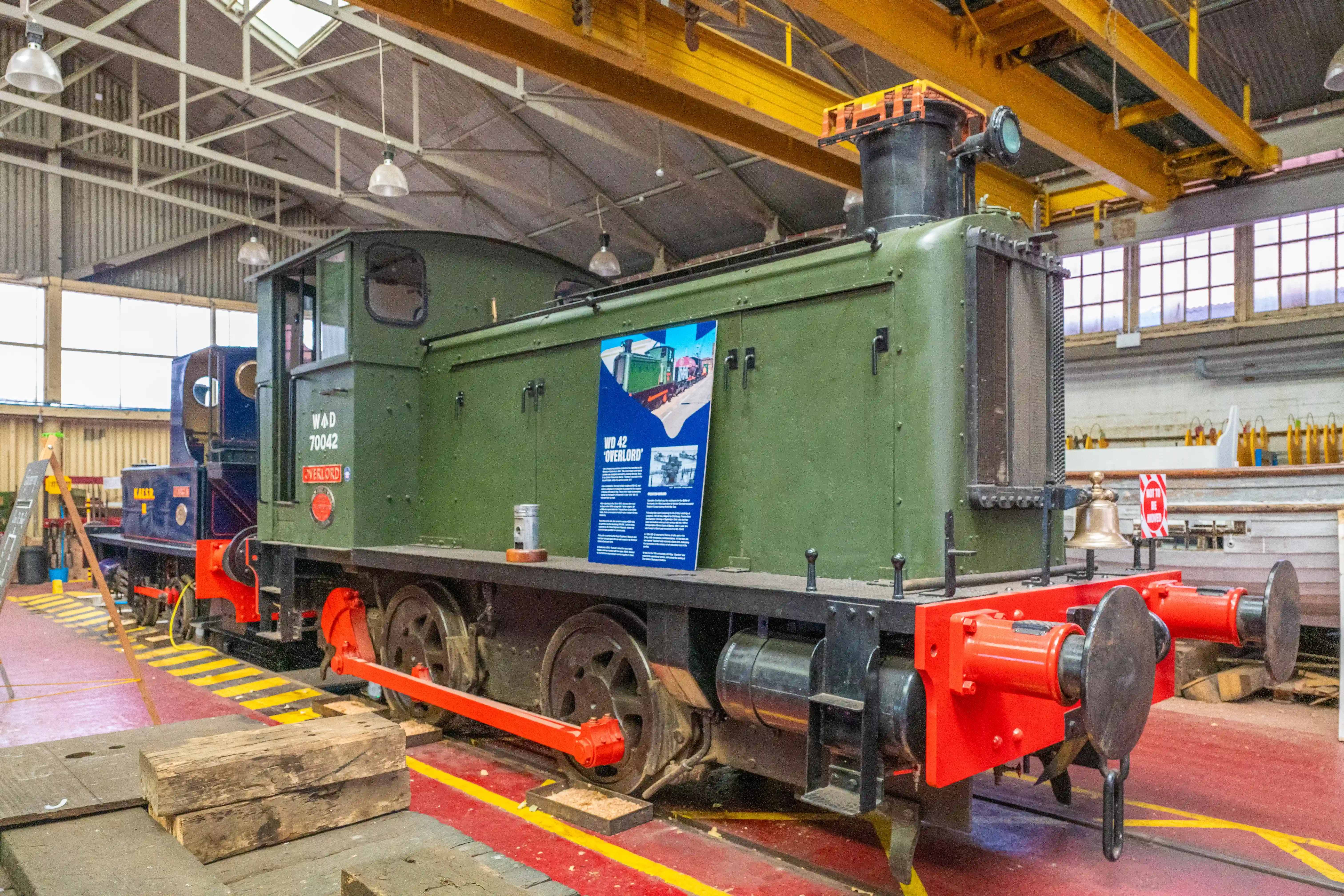 The image depicts a green locomotive, labeled 'Overlord' with the number W.D 70042, displayed in an indoor setting, likely a museum or workshop. The locomotive is accompanied by an information board detailing its history and specifications. The area is well-lit with industrial lighting and contains other railway equipment and machinery. The environment suggests a focus on preservation and education about historical trains.