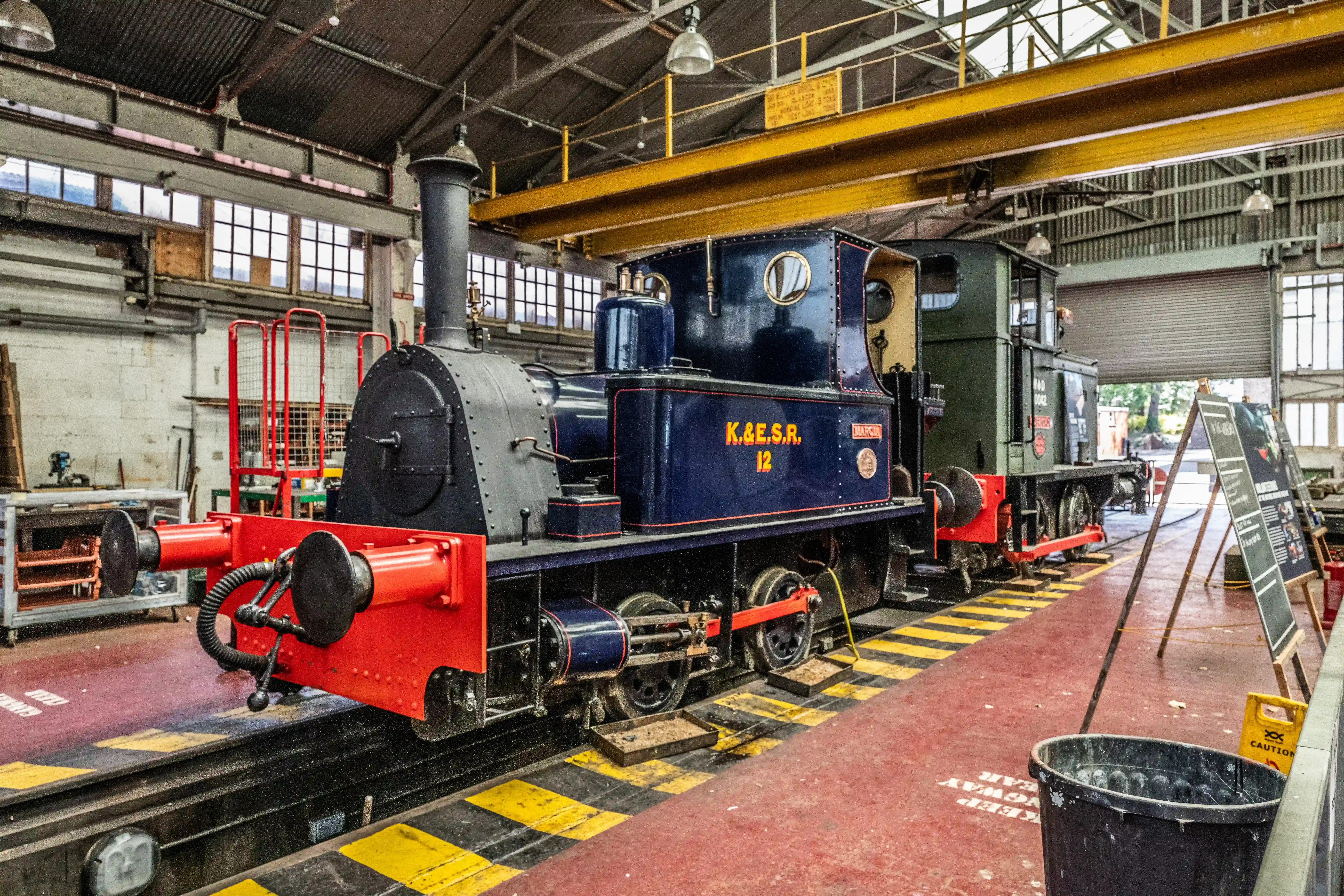 The image depicts an indoor workshop or maintenance facility housing several steam locomotives. The primary locomotive in the foreground is painted in dark blue and red, with the marking 'K&ESR 12' on its side. The facility is equipped with overhead cranes and various tools and equipment, indicating it is used for the repair and maintenance of trains. The area is well-lit with natural light coming through large windows, and there are safety signs and containers placed around.