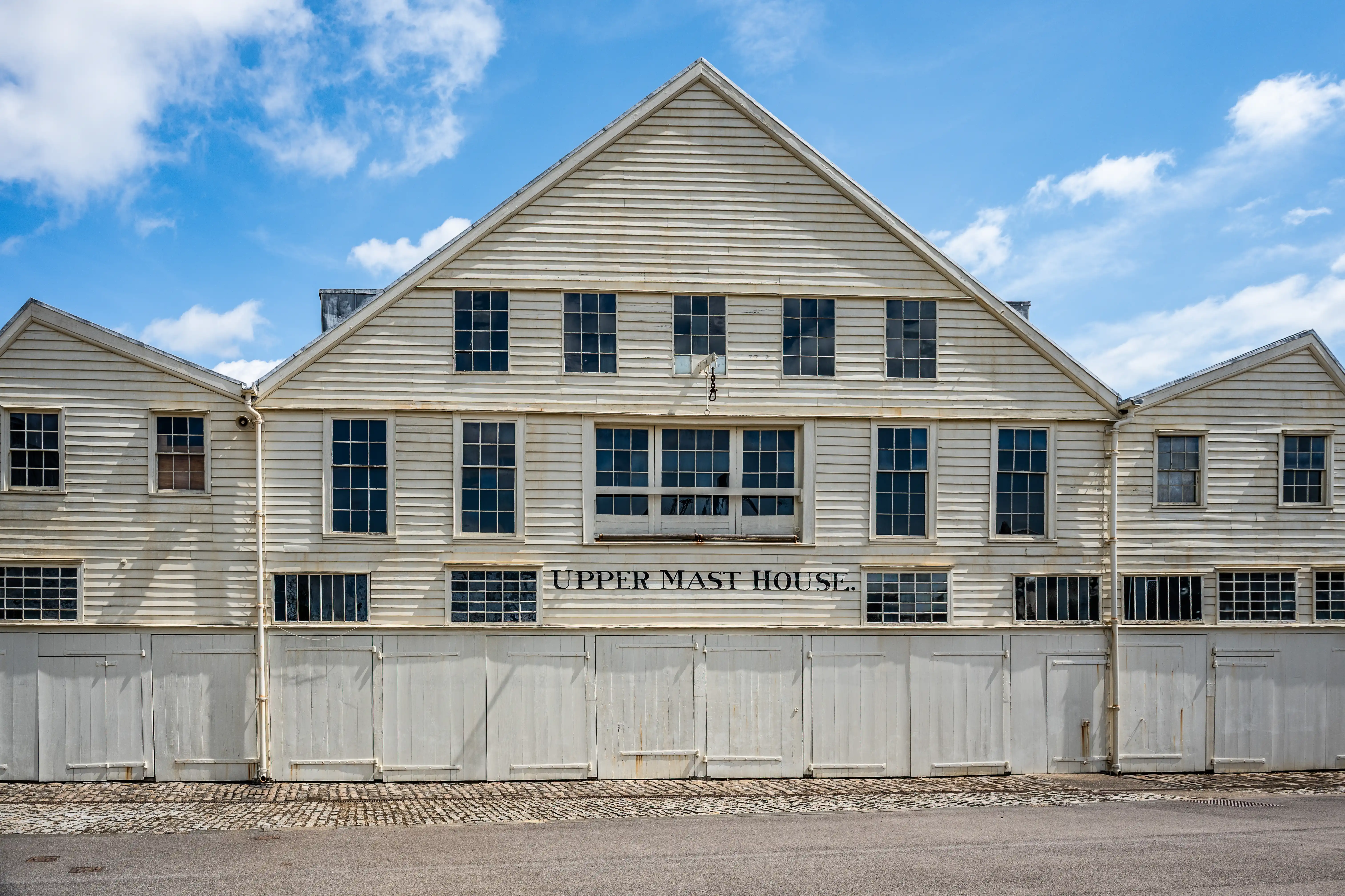 The image depicts a large, white, weathered building with a sign that reads 'Upper Mast House.' The structure has multiple large windows and doors on the ground level, and it appears to be part of a historical or industrial site. The building is situated on a paved area with a clear blue sky overhead.