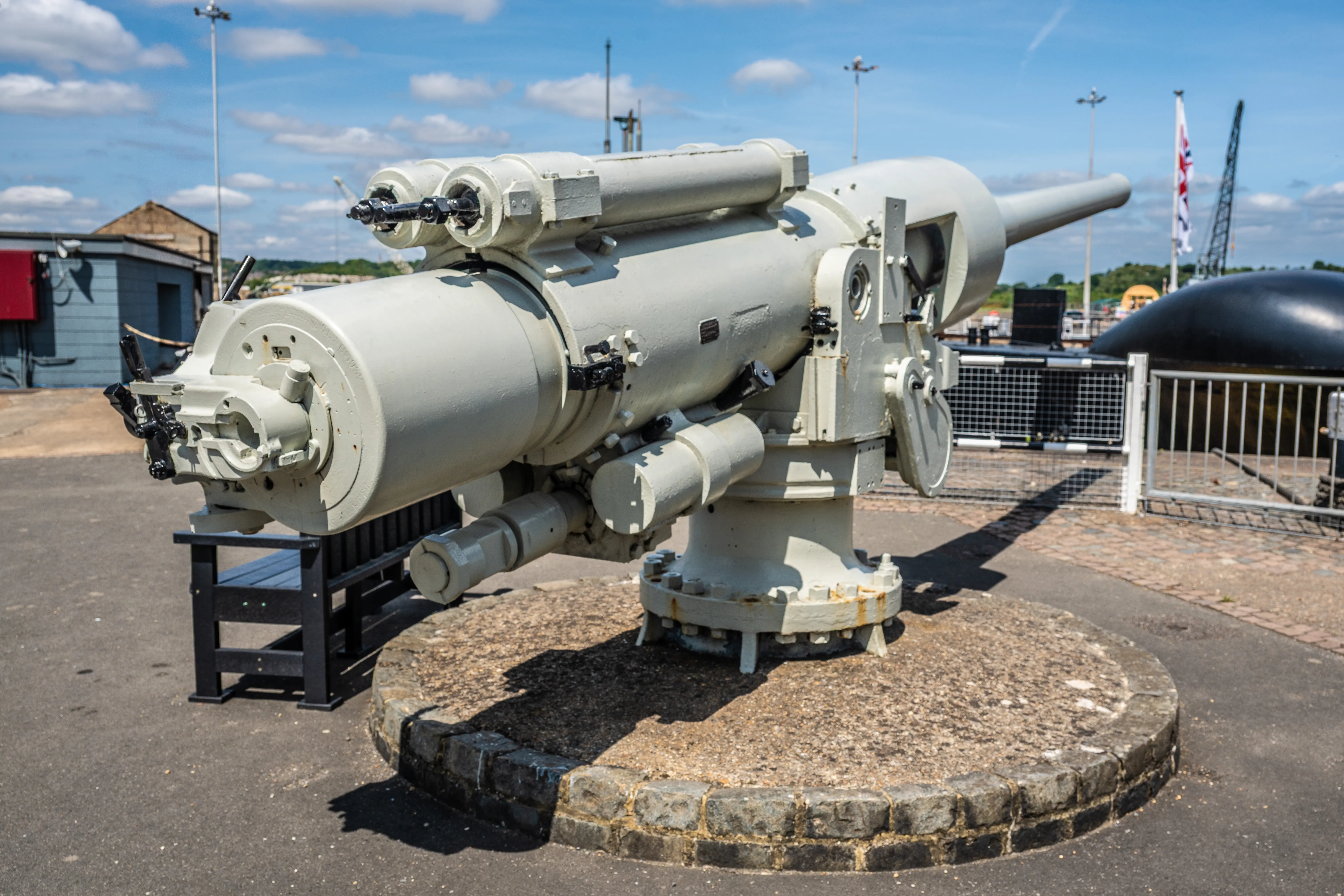 The image depicts a large, mounted naval gun, likely from a historical warship or submarine. The gun is displayed outdoors on a concrete platform with a circular stone border. It is painted in a light color, possibly white or gray, and is equipped with various mechanical components. The background includes industrial structures, cranes, and a clear blue sky.