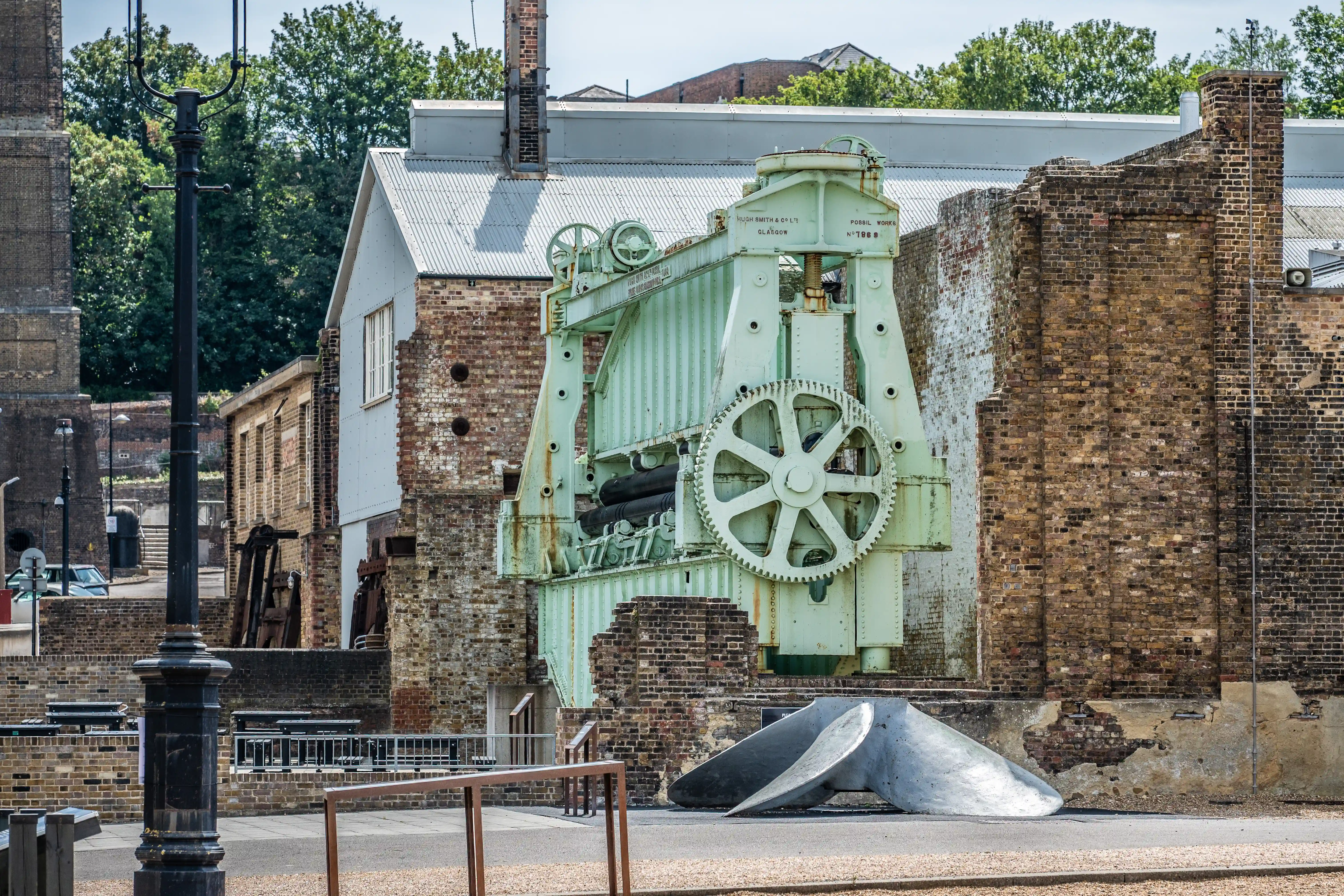 The image depicts an old industrial site featuring a large, green, mechanical hoist or winding gear prominently displayed in the center. The machinery is set against a backdrop of aged brick buildings, some of which are partially ruined. There is a modern lamp post in the foreground, and the scene is surrounded by a mix of historical and slightly modernized structures. The overall setting suggests a preserved historical area, possibly a museum or heritage site.
