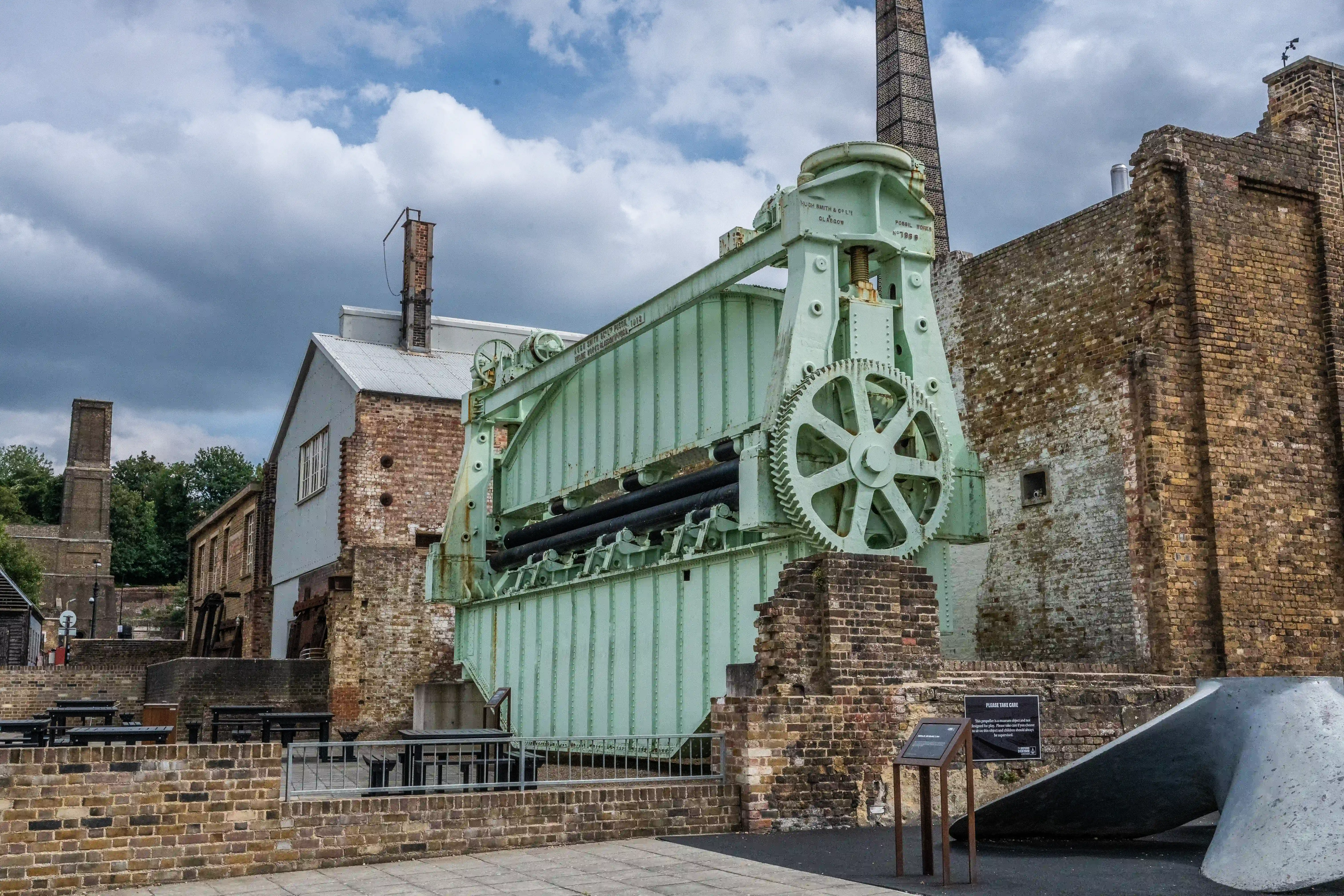 The image depicts an old industrial site with a prominent piece of machinery, likely a steam-powered beam engine, situated among brick buildings. The machinery is painted green and appears to be well-preserved, suggesting it might be part of a historical exhibit or museum. The surrounding buildings are constructed from brick and show signs of age and wear. The sky is partly cloudy, adding to the historical ambiance of the scene. There are informational plaques and seating areas, indicating that the site is accessible to visitors.