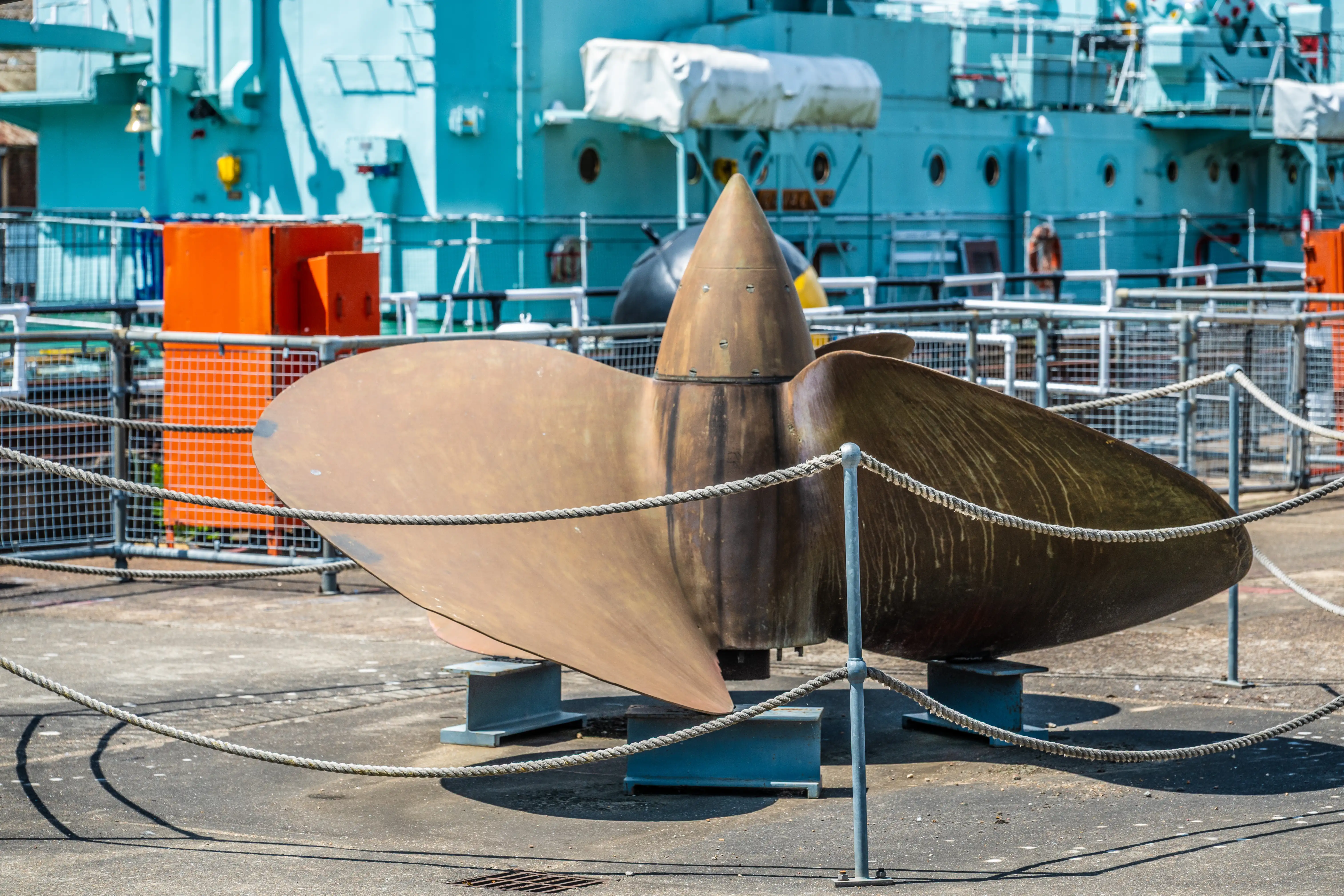 The image depicts a large ship propeller displayed on a dock. The propeller is cordoned off with ropes and stands on a metal support. In the background, there is a large ship with a light blue hull and various equipment. The scene appears to be part of a maritime or naval setting, possibly a shipyard or museum.