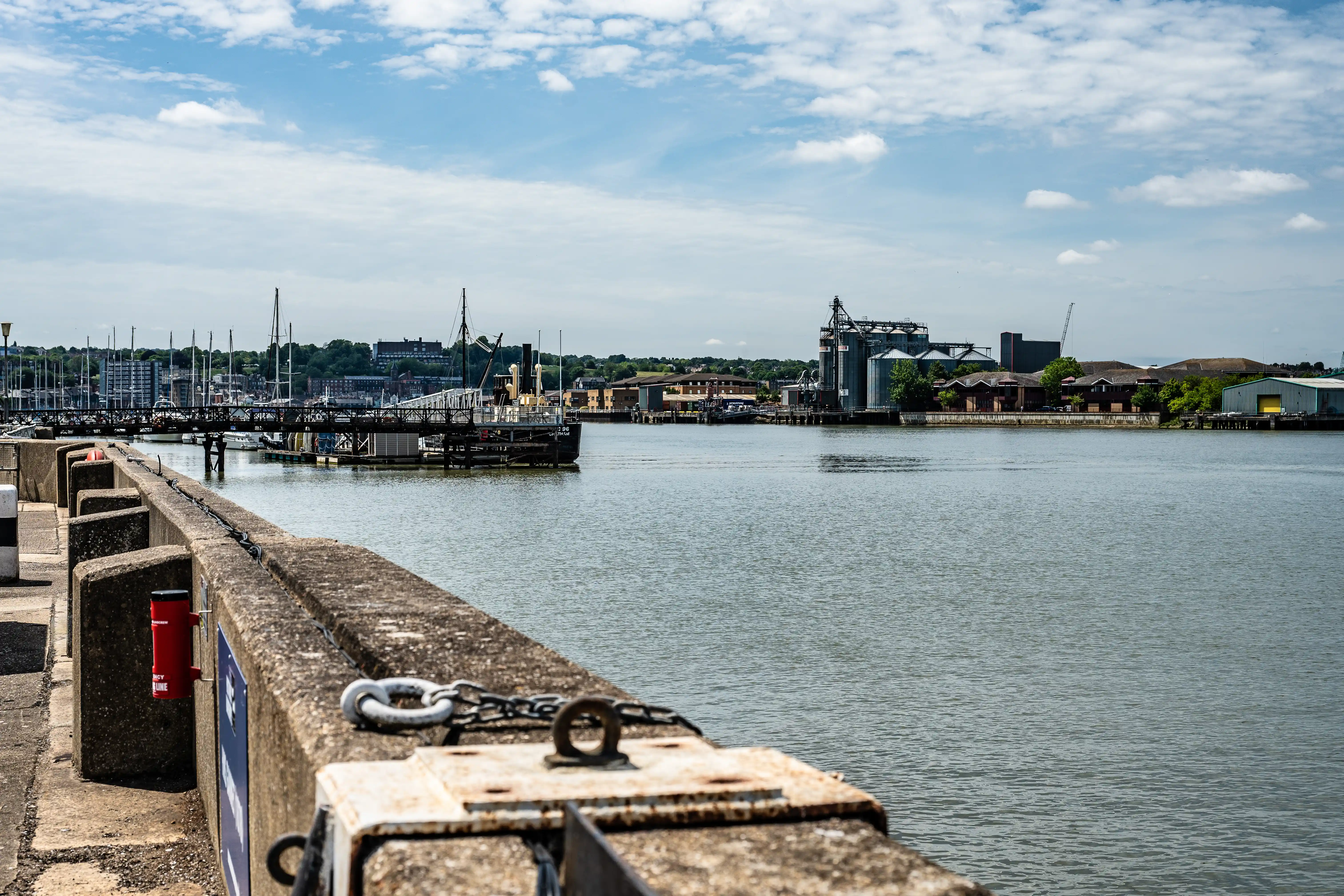 The image depicts a waterfront scene with a calm body of water, likely a river or harbor. There is a concrete walkway with a metal railing in the foreground. In the background, industrial buildings and silos are visible, along with some greenery and a few boats docked at piers. The sky is mostly clear with some scattered clouds, indicating a bright and sunny day.