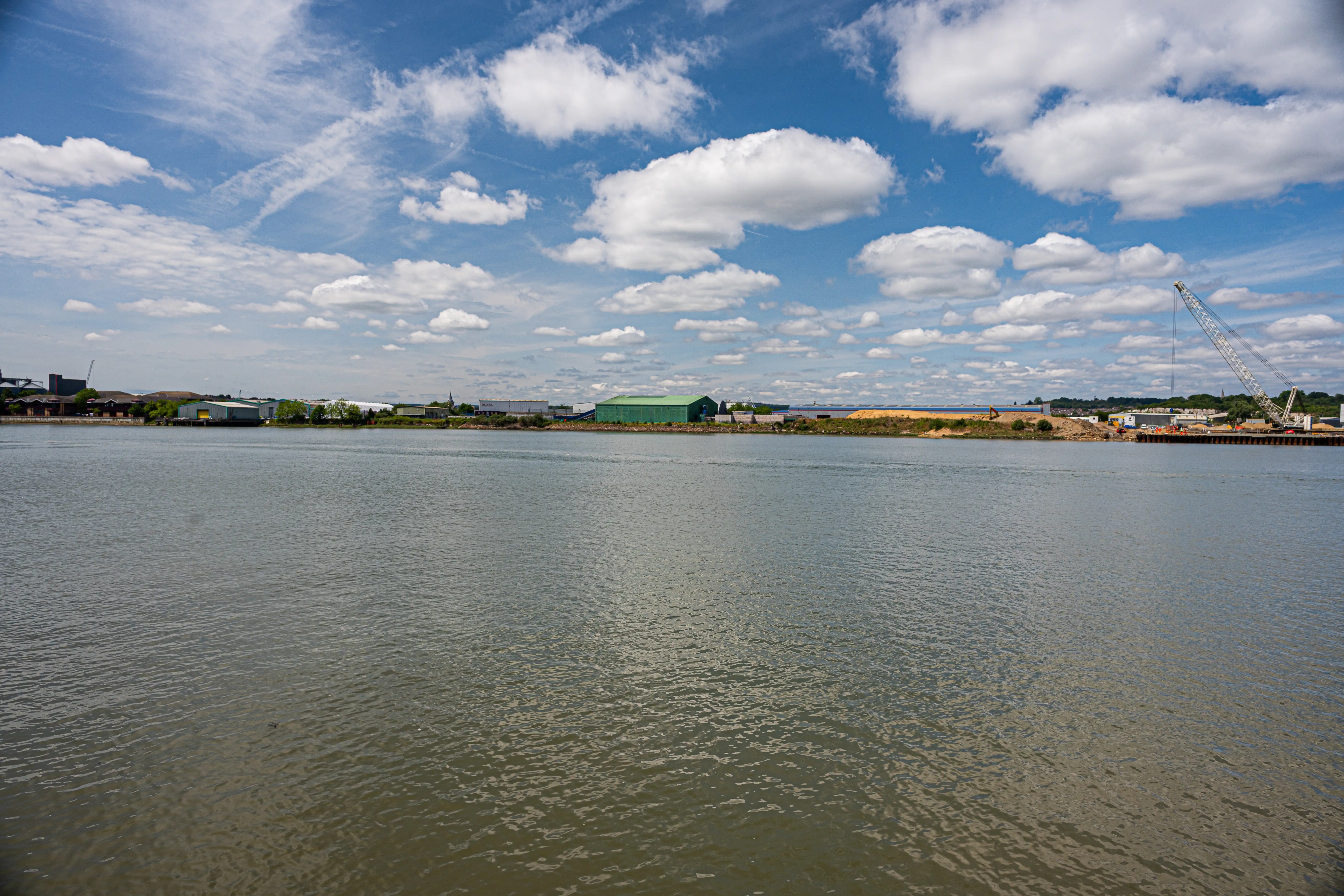 The image depicts a waterfront scene with a calm body of water, likely a river or harbor. There is a concrete walkway with a metal railing in the foreground. In the background, industrial buildings and silos are visible, along with some greenery and a few boats docked at piers. The sky is mostly clear with some scattered clouds, indicating a bright and sunny day.