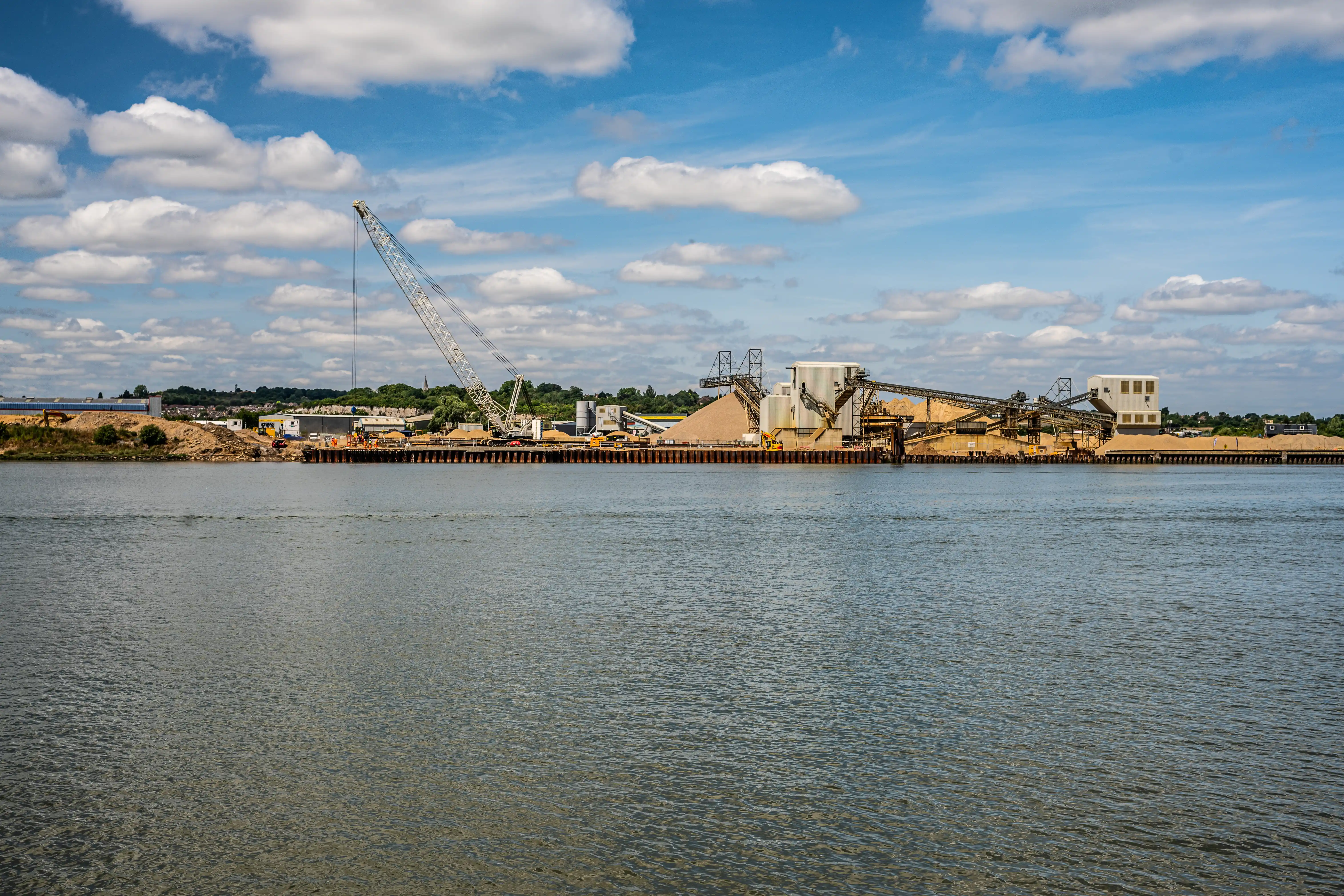 The image depicts an industrial site located by a body of water. The site features a large crane, conveyor belts, and several buildings, likely used for processing or storing materials. There are substantial piles of what appears to be sand or aggregate materials. The scene is set under a partly cloudy sky, suggesting a typical day at an industrial facility, possibly involved in construction or shipping activities.