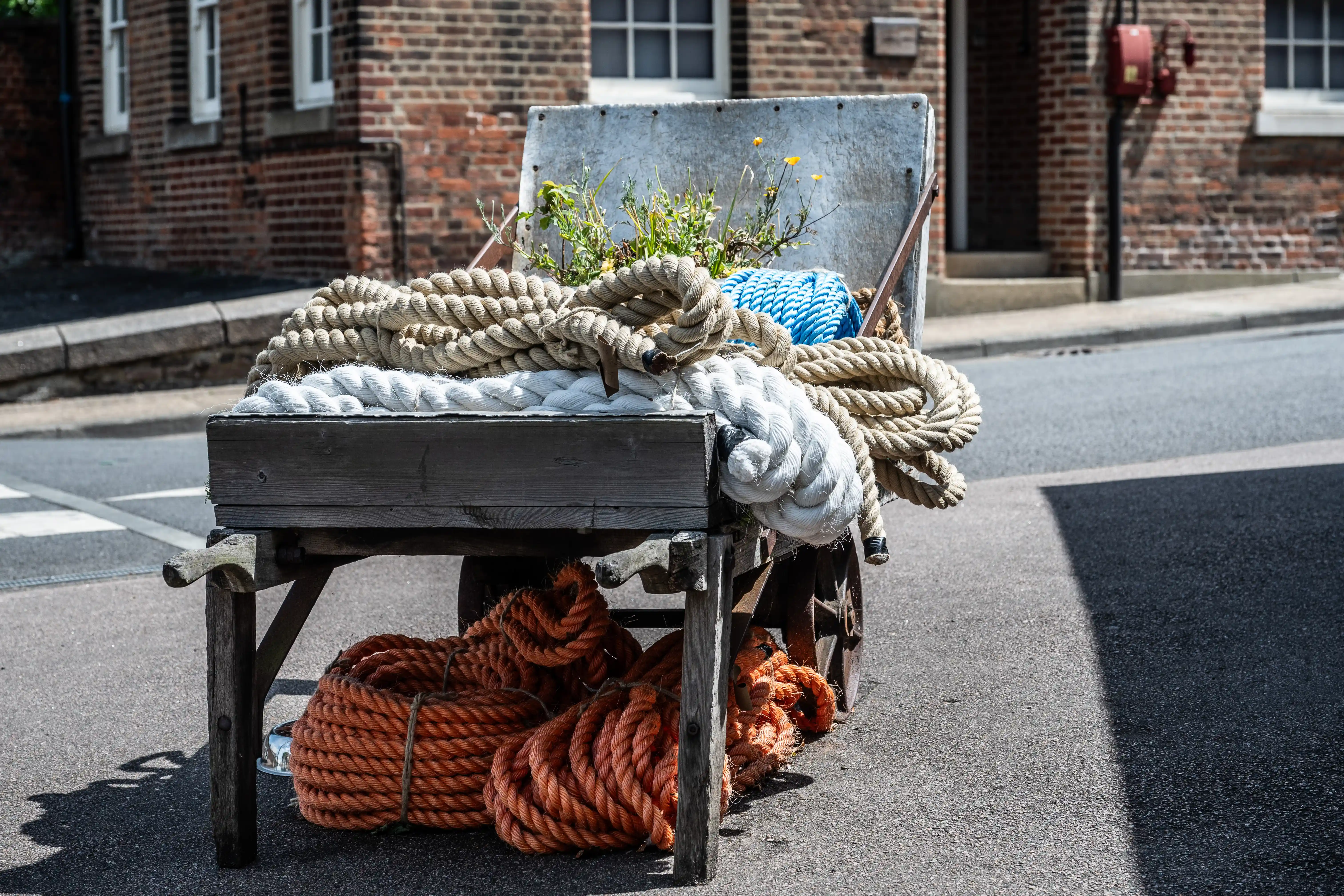 The image depicts a wooden cart positioned on a street. The cart is loaded with various types of ropes, including thick white ropes on top and orange ropes on the bottom shelf. There is also a blue net visible on the cart. The cart is situated in front of a brick building with white-framed windows.