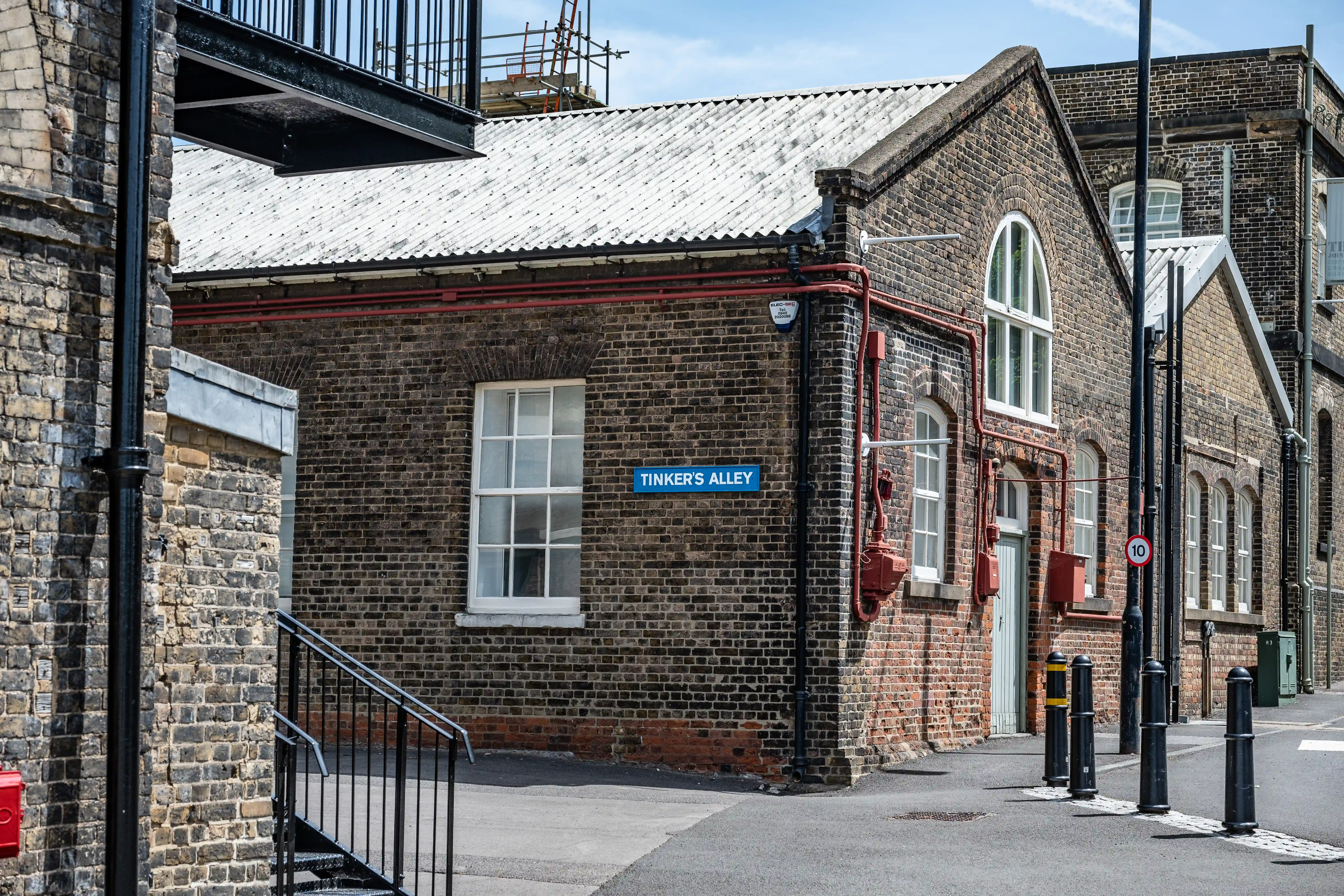 The image depicts a street view of a brick building identified as 'Tinker's Alley.' The building has a corrugated metal roof and several windows with white frames. Red pipes run along the exterior walls, and there are black bollards lining the street. The area appears to be part of an older, industrial or historical section of a town or city.