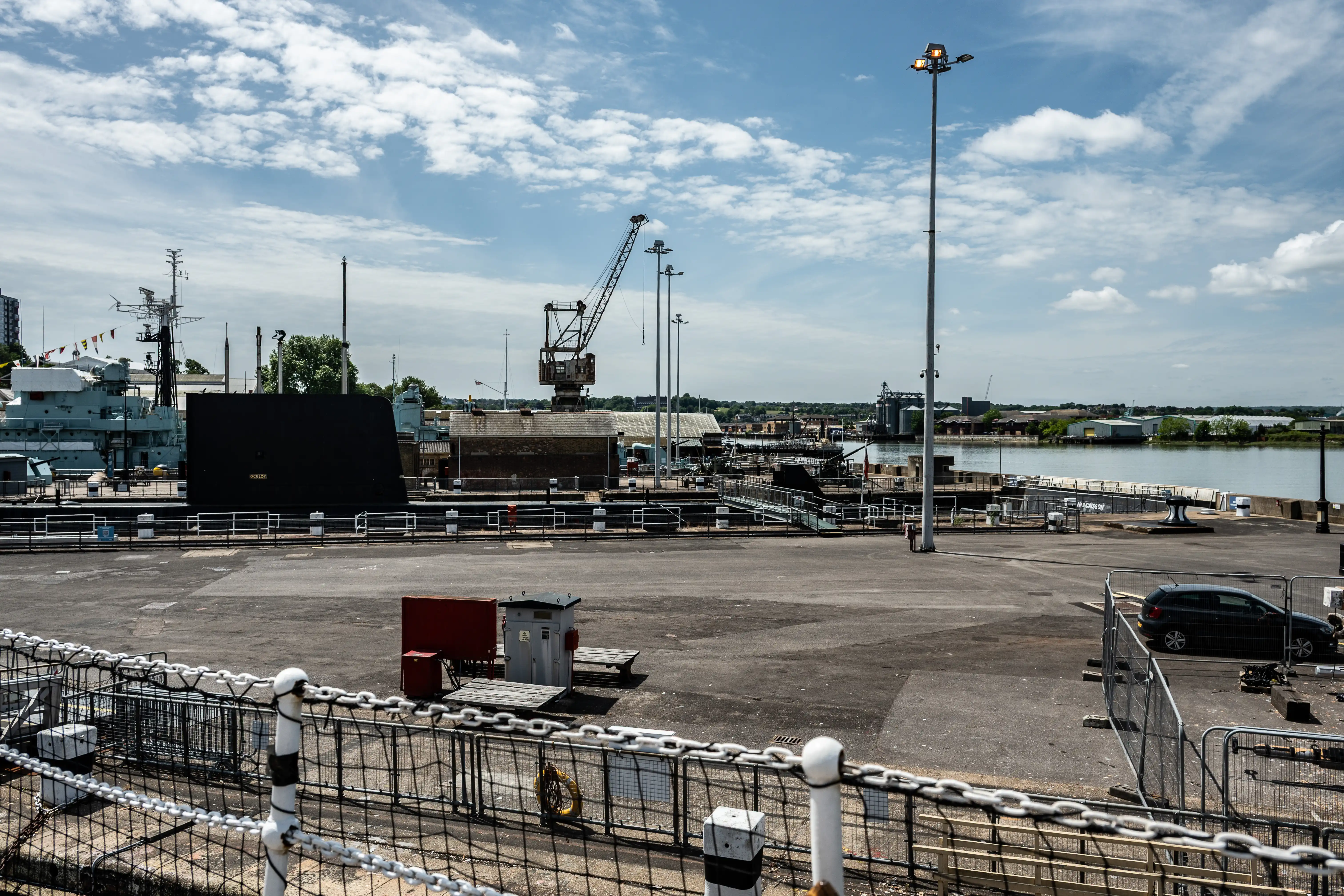 The image depicts a dockyard scene. There is a large naval ship docked on the left side, with a crane and other industrial structures visible in the background. The dockyard area is spacious, with a few vehicles and equipment scattered around. The sky is clear with some clouds, and the overall setting appears to be a maritime or naval base.