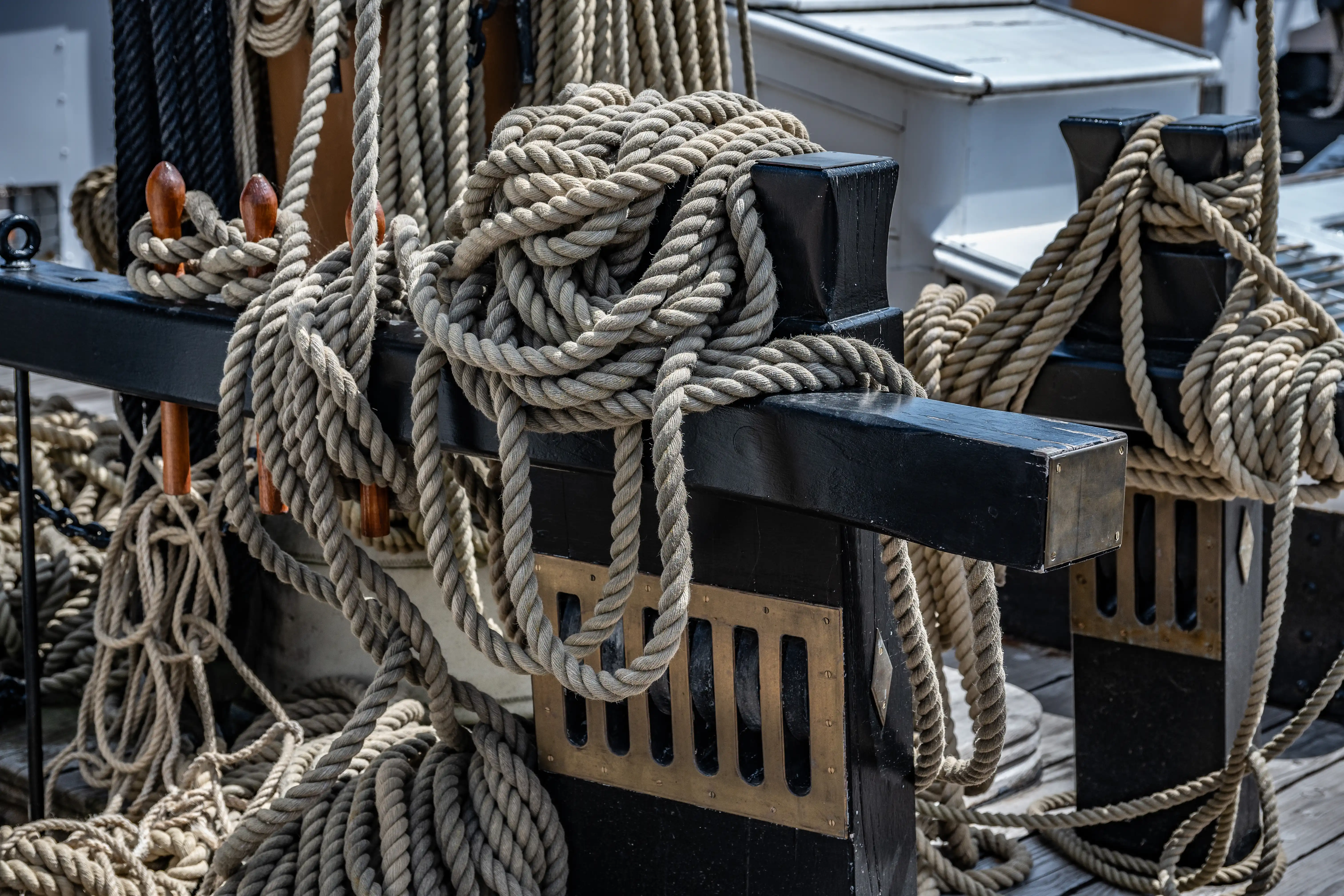 The image depicts a close-up view of a section of a ship's deck, featuring intricately tied ropes and knots secured to black metal cleats. The ropes are thick and appear well-used, suggesting the ship is actively in use. The background includes additional ropes and parts of the ship's structure, indicating a maritime setting. The focus on the knots highlights traditional nautical skills and craftsmanship.