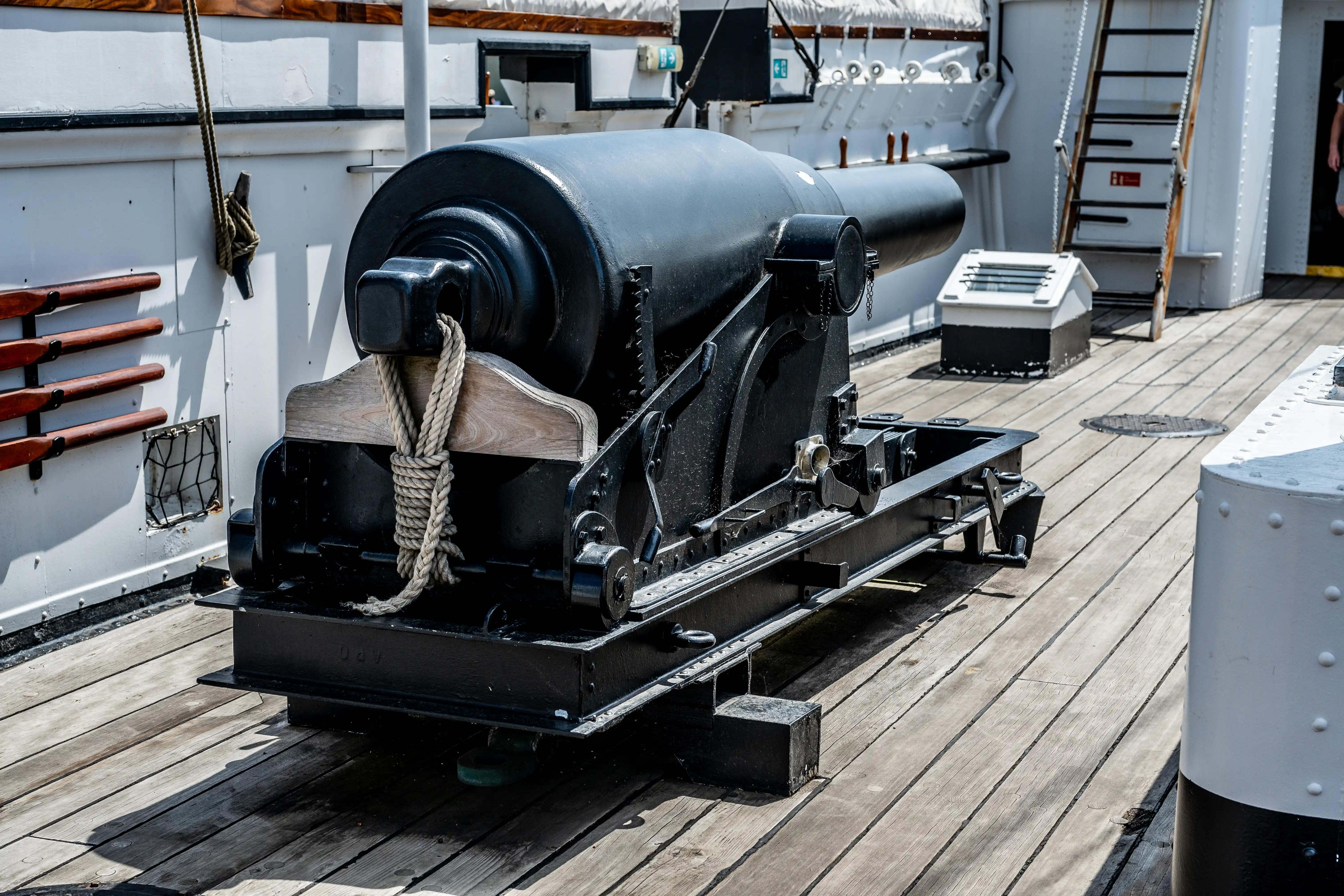 he image depicts a historical naval cannon mounted on a wooden deck. The cannon is secured with ropes and positioned on a carriage, indicating it is likely part of a historical ship or a museum exhibit. The surrounding area includes parts of the ship's structure, such as railings and ladders, and the deck is made of wooden planks. The setting suggests a maritime or naval theme, possibly from a bygone era of sailing ships and naval warfare.
