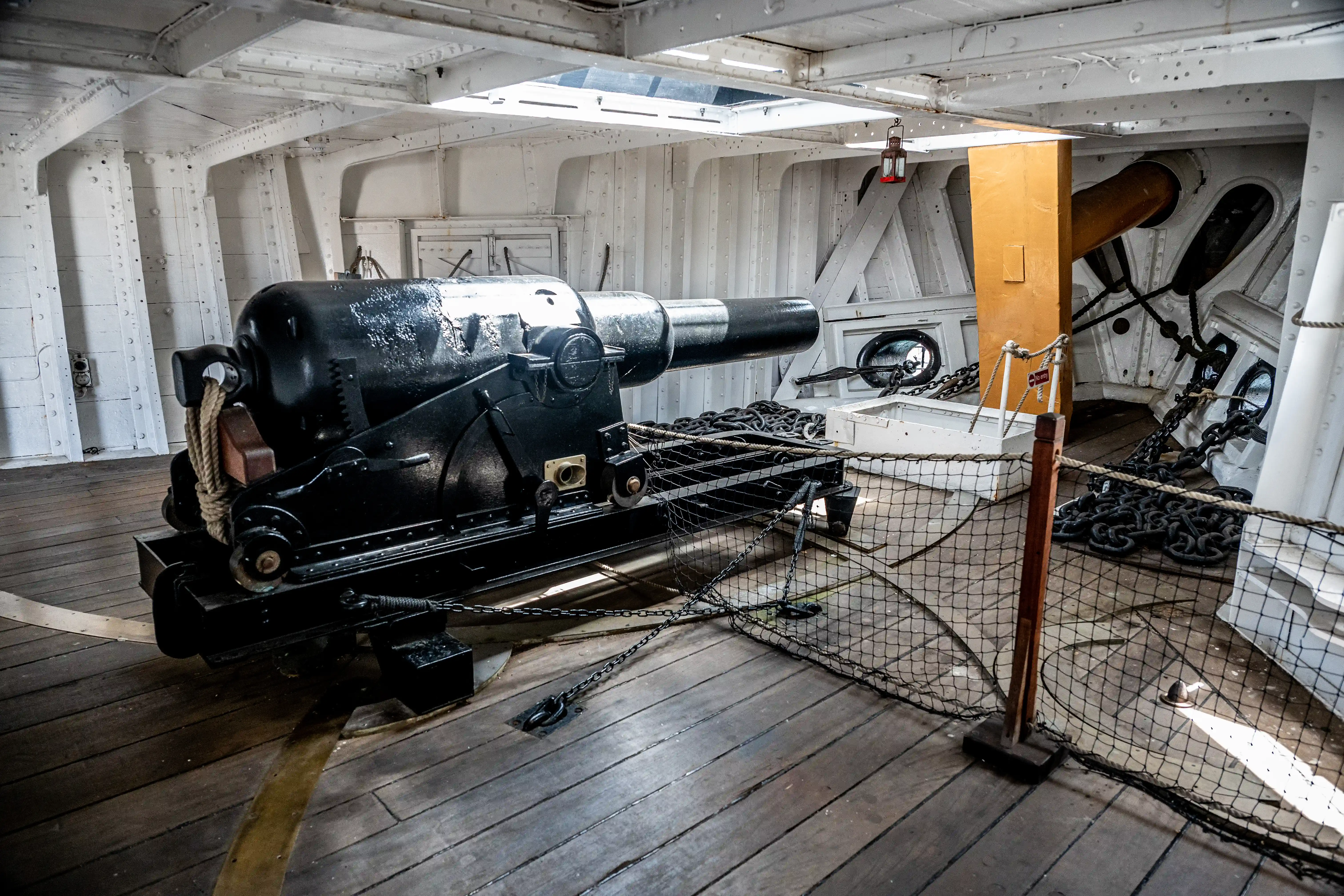 The image shows the interior of a historical naval ship, featuring a large black cannon mounted on a wooden deck. The cannon is secured with chains and ropes, and the surrounding area is equipped with various nautical elements such as a yellow post, additional chains, and a net. The walls and ceiling are painted white, and there are skylights allowing natural light to enter. The overall setting suggests a well-preserved or restored ship, likely from a museum or historical site.