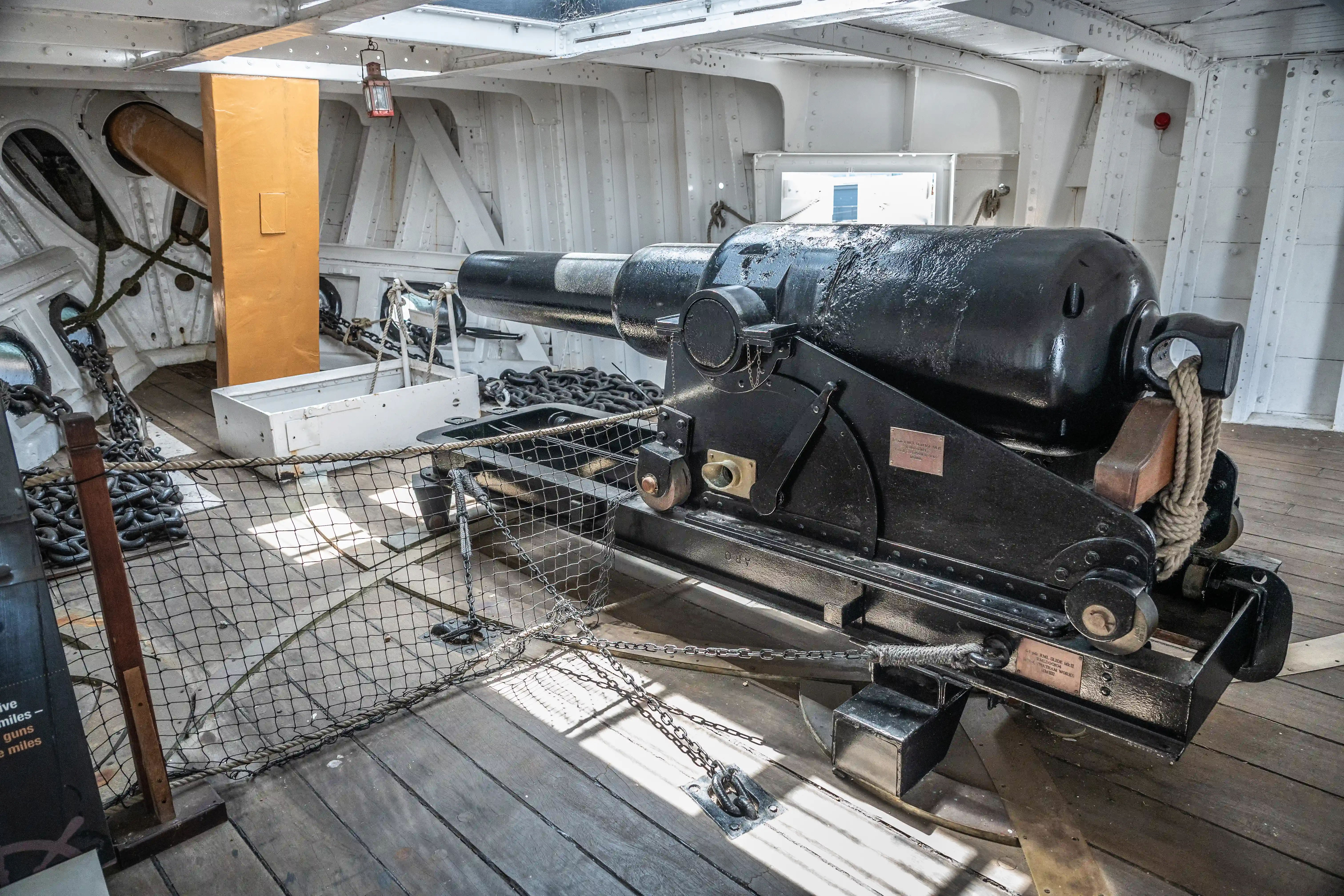 The image depicts the interior of a historical naval ship, showcasing a large cannon mounted on a wooden deck. The cannon is secured with chains and ropes, and there are various other naval elements visible, such as additional cannons, metal structures, and informational plaques. The setting appears to be part of a museum or historical exhibit, highlighting maritime artillery and ship design from a past era.