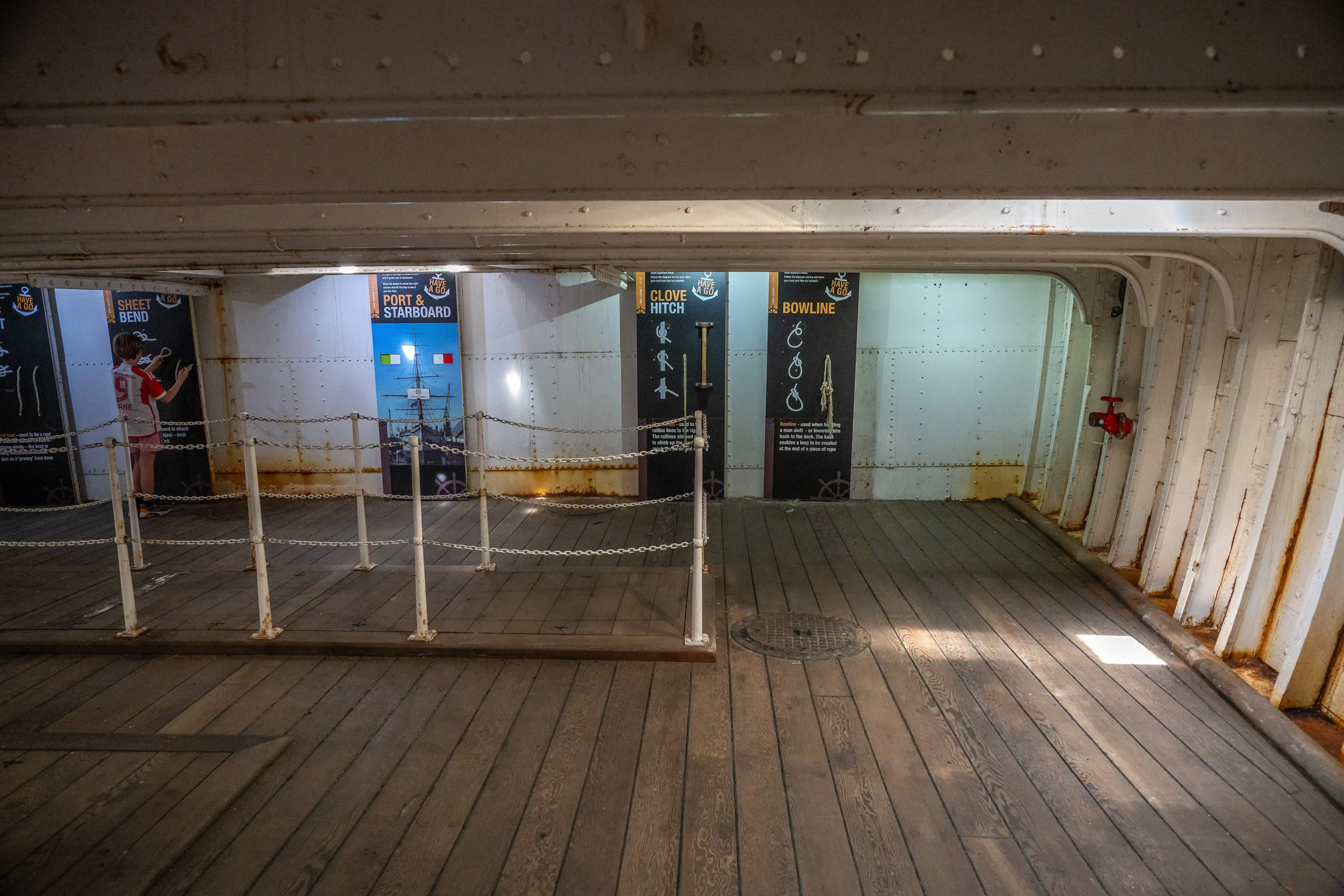 The image depicts an interior section of a ship with informational posters on the walls. The posters provide instructions on various nautical knots and procedures such as the Sheet Bend, Port & Starboard, Clove Hitch, and Bowline. The area is cordoned off with chains and has a wooden floor. A person is visible in the background, seemingly interacting with one of the posters.