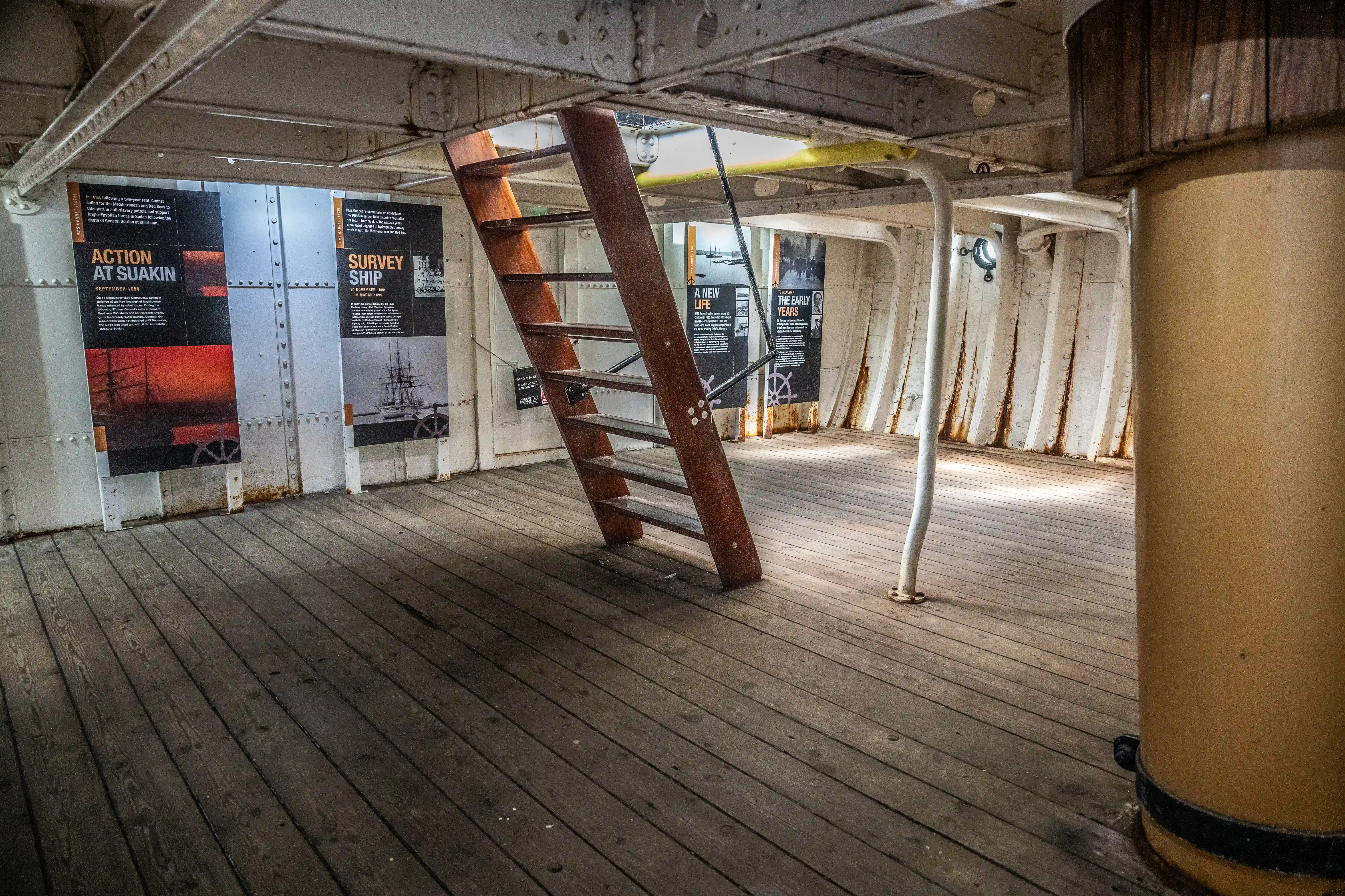 The image depicts an interior section of a historical ship, likely a museum exhibit. Informational posters line the walls, detailing events such as 'Action at Suakin' and 'Survey Ship.' A wooden ladder is prominently positioned in the center, leading to another deck. The space is characterized by wooden flooring and metal structural elements, giving a sense of the ship's construction and historical context.