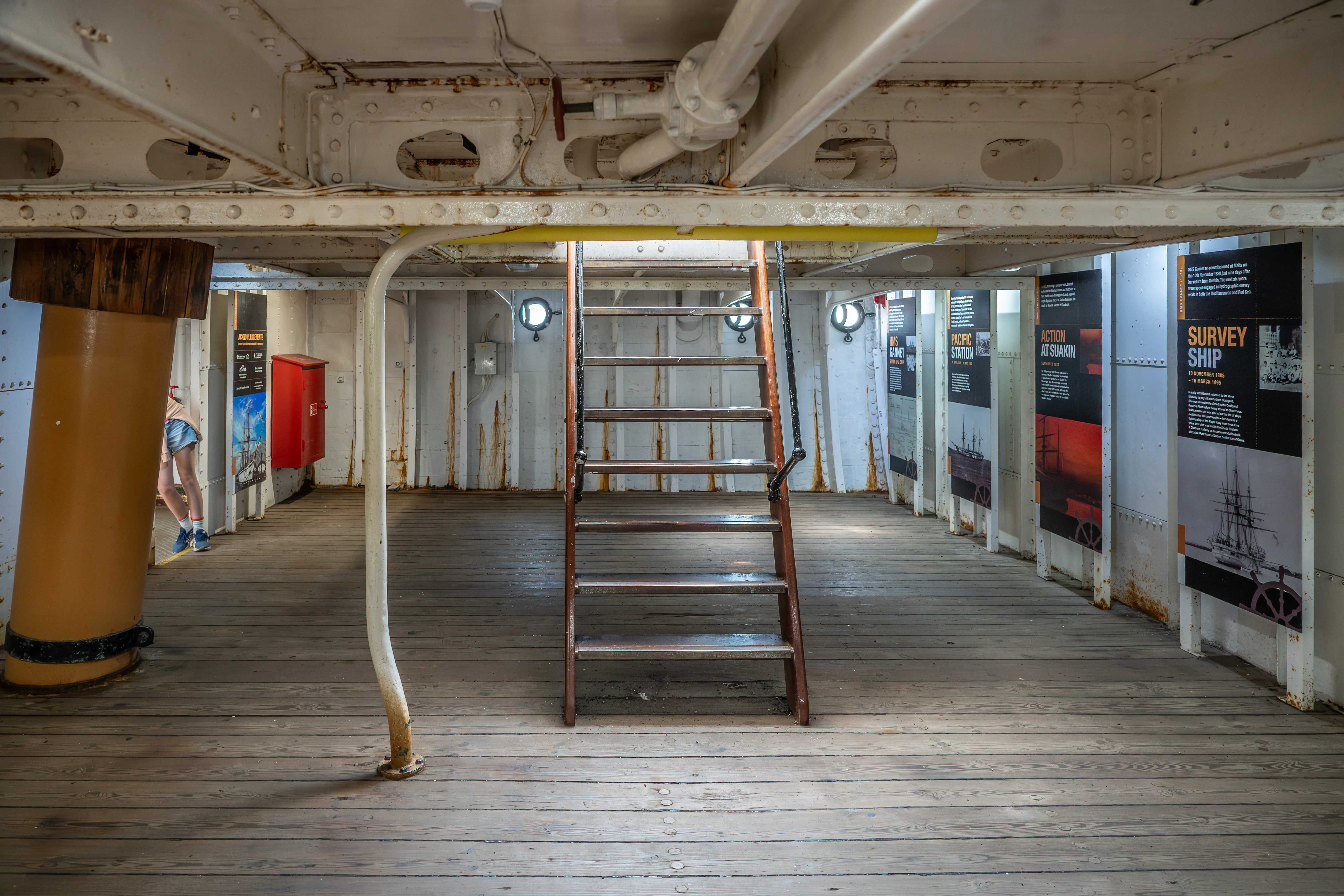 The image depicts the interior of a historical ship, likely a museum or exhibit. The area features a wooden deck, metal structures, and informational panels detailing various aspects of the ship's history and operations. A ladder leads to another deck, and there are various pipes and structural elements visible. A person is seen walking through the exhibit, and there is a red fire safety box mounted on the wall.