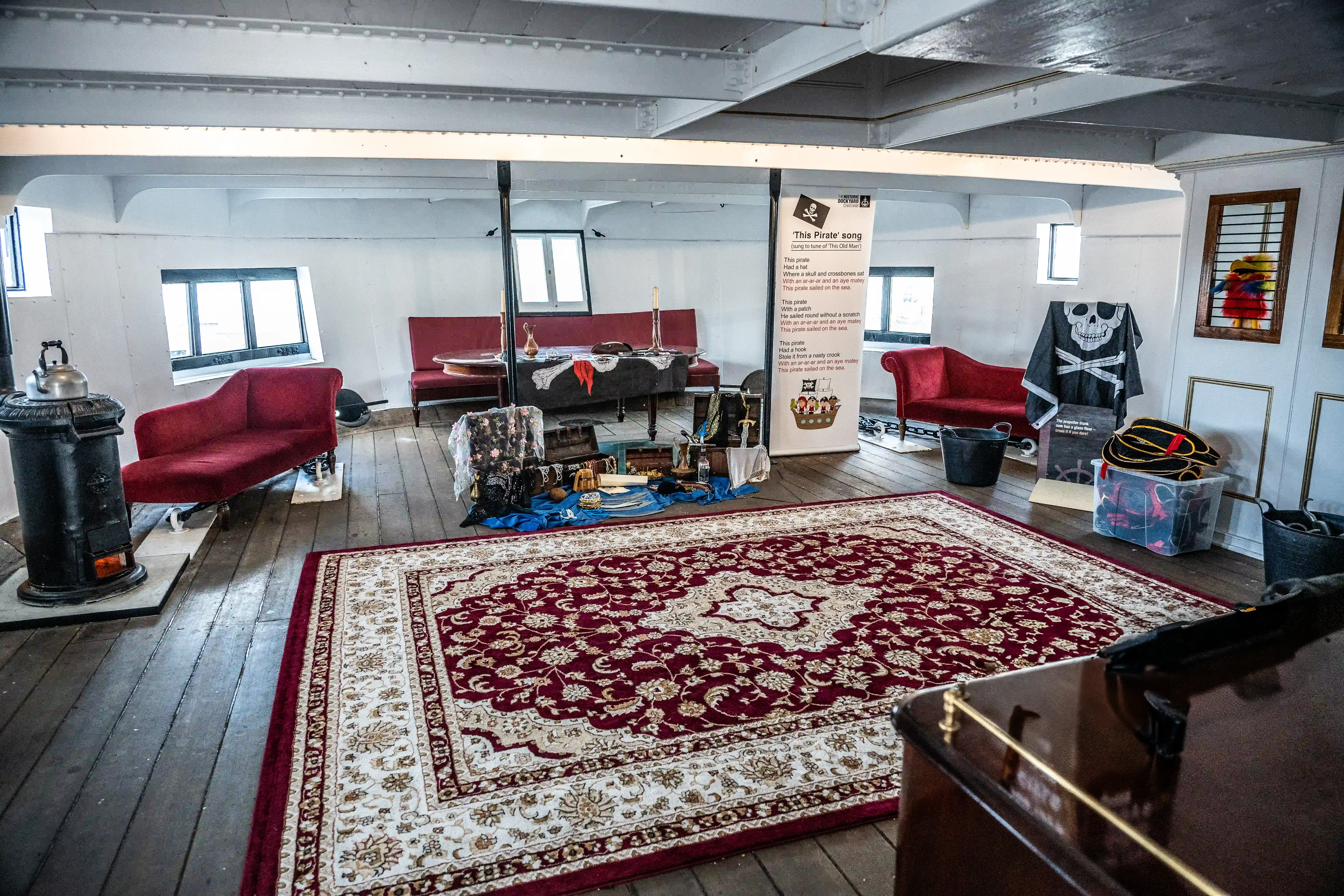 The image depicts an interior space designed to resemble a pirate-themed room. It features vintage furniture, including red sofas and a large patterned rug. There is a display of pirate-related items and a sign explaining the theme song 'This Pirate.' The room also includes a stove and various pirate decorations, such as a skull and crossbones flag.