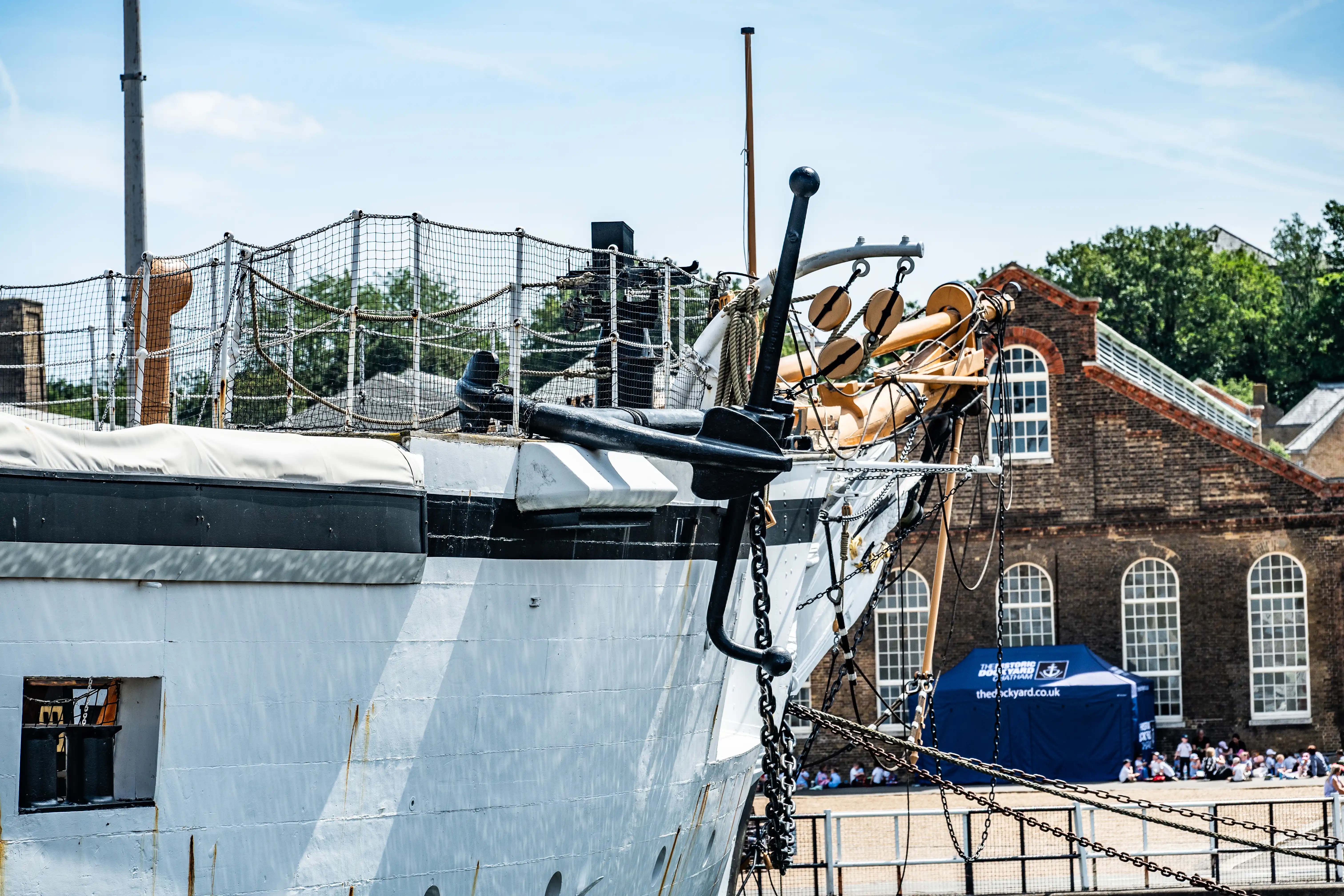 The image depicts the bow of a historical ship, showcasing its anchor and various rigging elements. The ship is docked near a brick building, with a crowd of people visible in the background. There is a blue tent with text, indicating some sort of event or exhibition. The ship's structure and details suggest it might be part of a maritime museum or historical site.