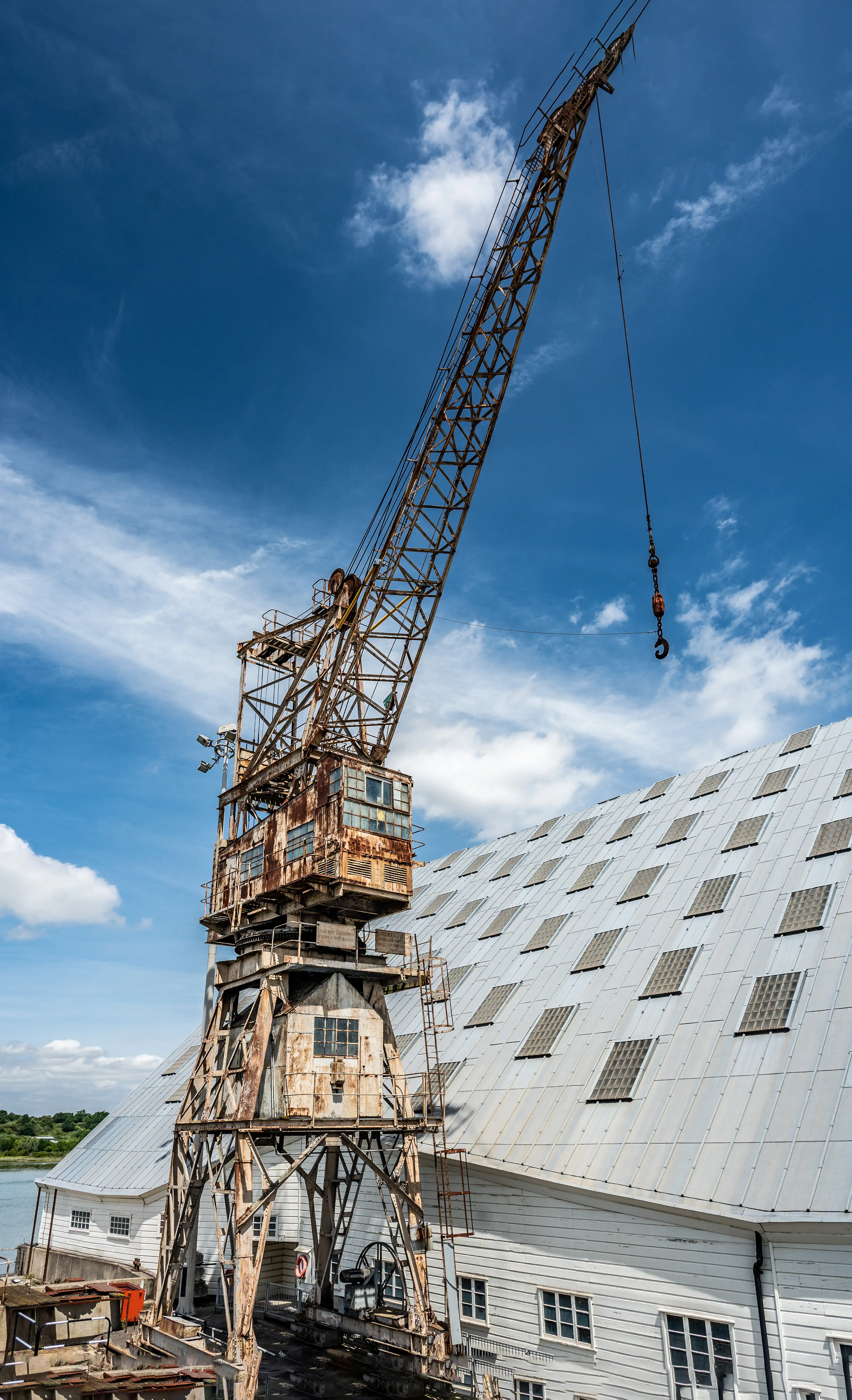 The image depicts an old, rusty industrial crane situated next to a large building with a sloped metal roof. The crane appears to be weathered and aged, showing signs of rust and wear. The building adjacent to the crane has a modern, industrial look with a series of windows. The sky above is mostly clear with a few scattered clouds, indicating a bright day. The scene suggests an industrial or dockyard setting, possibly used for loading and unloading heavy materials.