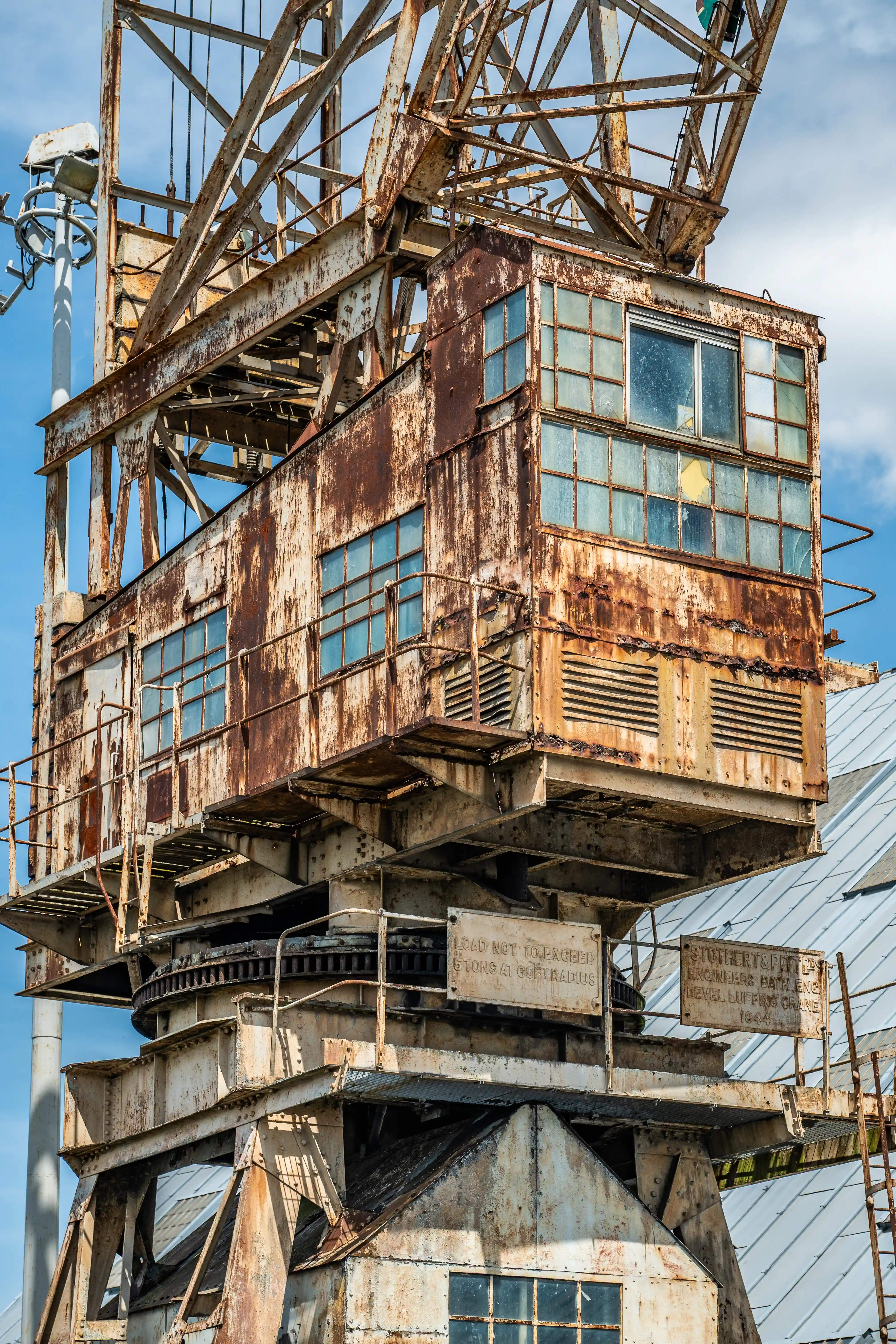 The image shows an old, rusted industrial structure, likely a part of a mining or construction site. The structure features a large, elevated cabin with multiple windows and a staircase leading to it. There are signs of significant wear and corrosion on the metal, indicating age and exposure to the elements. A sign on the structure reads a caution about load limits and credits engineers from 1934.