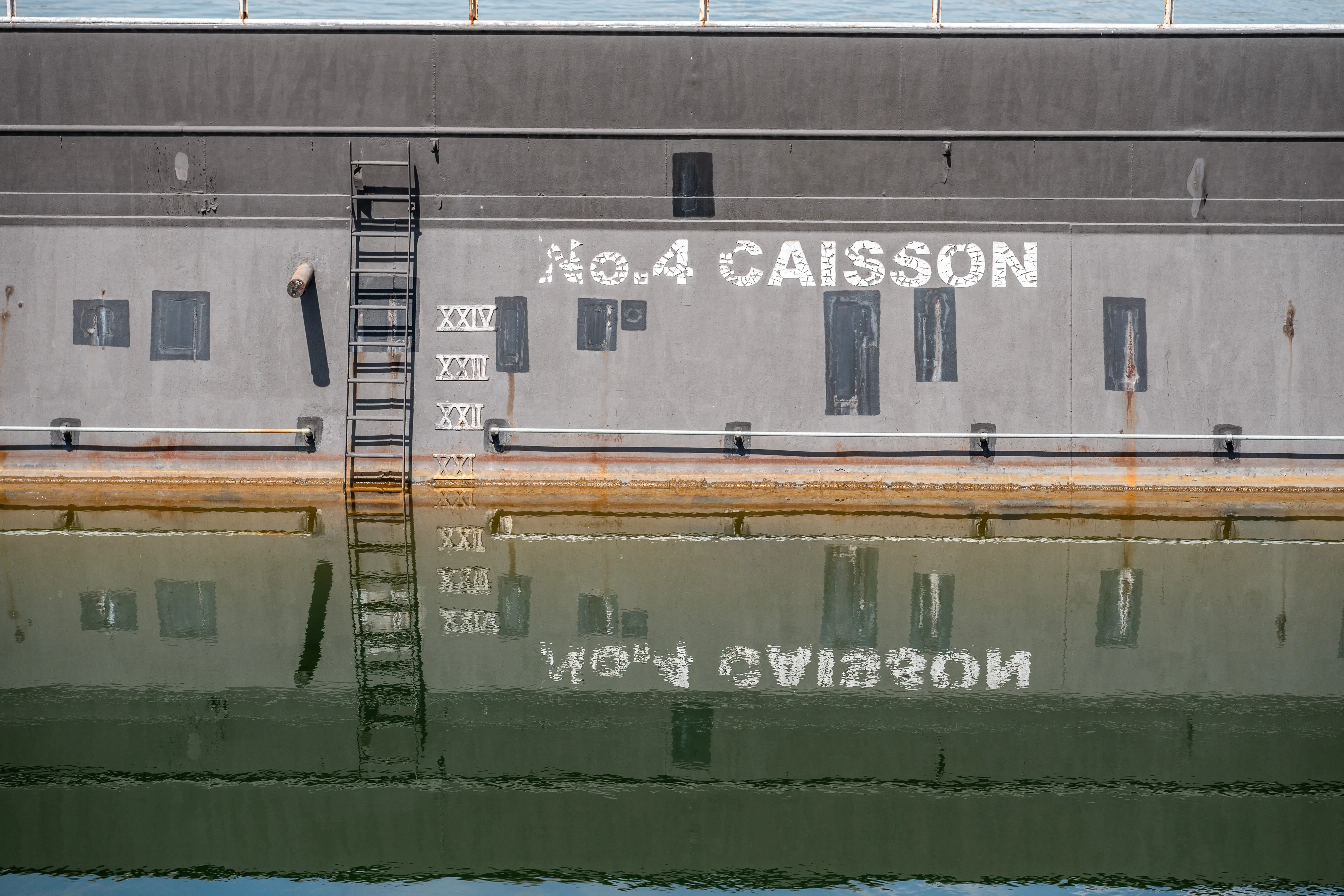 The image shows a section of a large water structure labeled 'No. 4 Caisson.' The structure has several windows and a ladder attached to its side. Roman numerals are marked on the wall, and the text on the structure is reflected in the calm water below.