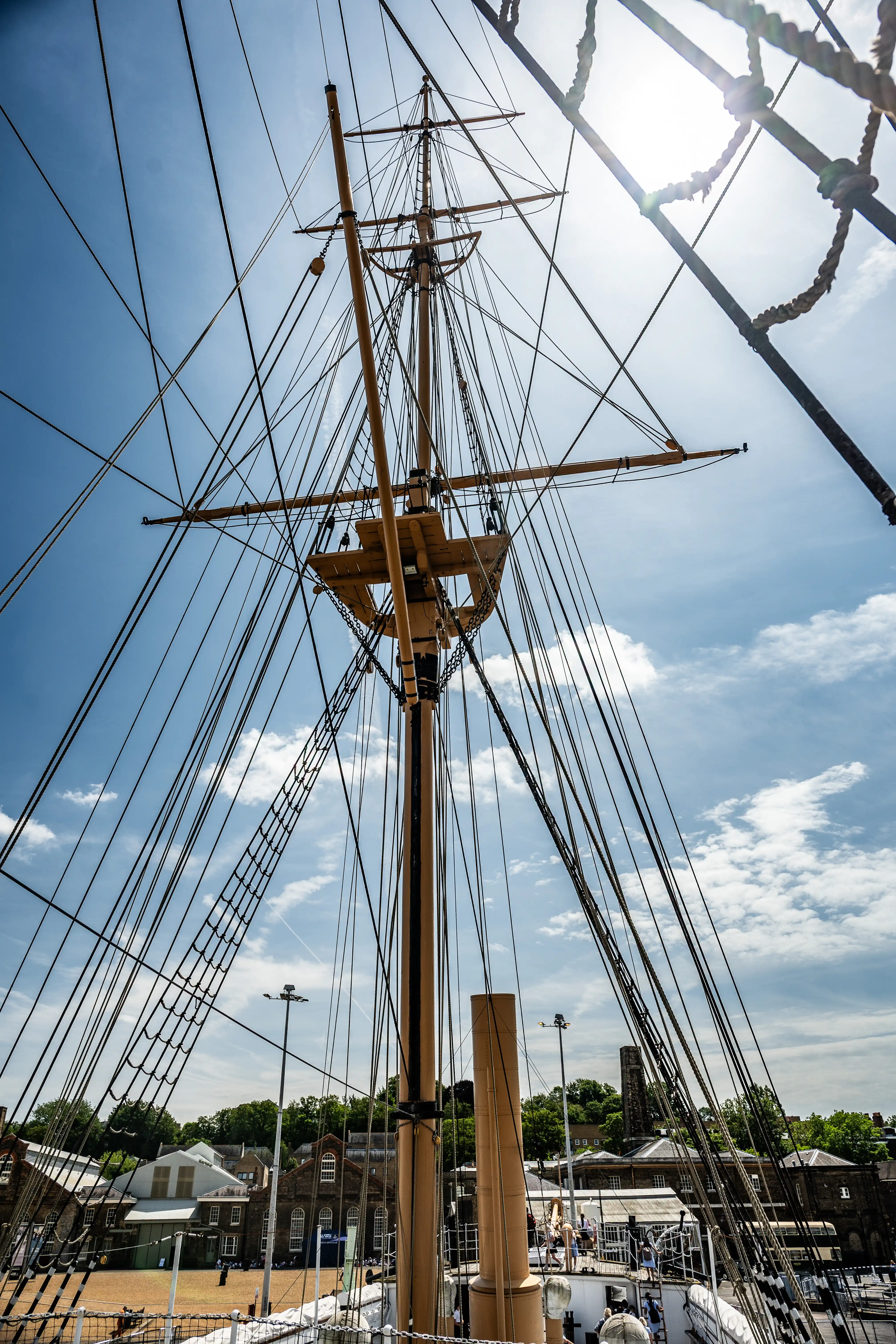 The image depicts the mast and rigging of a tall ship, with numerous ropes and sails extending from the central mast. The perspective is from the deck looking upwards towards the sky, showcasing the complexity and intricacy of the ship's rigging. In the background, there are buildings and greenery, suggesting the ship is docked in a harbor or port area. The sky is partly cloudy, adding to the dramatic effect of the scene.