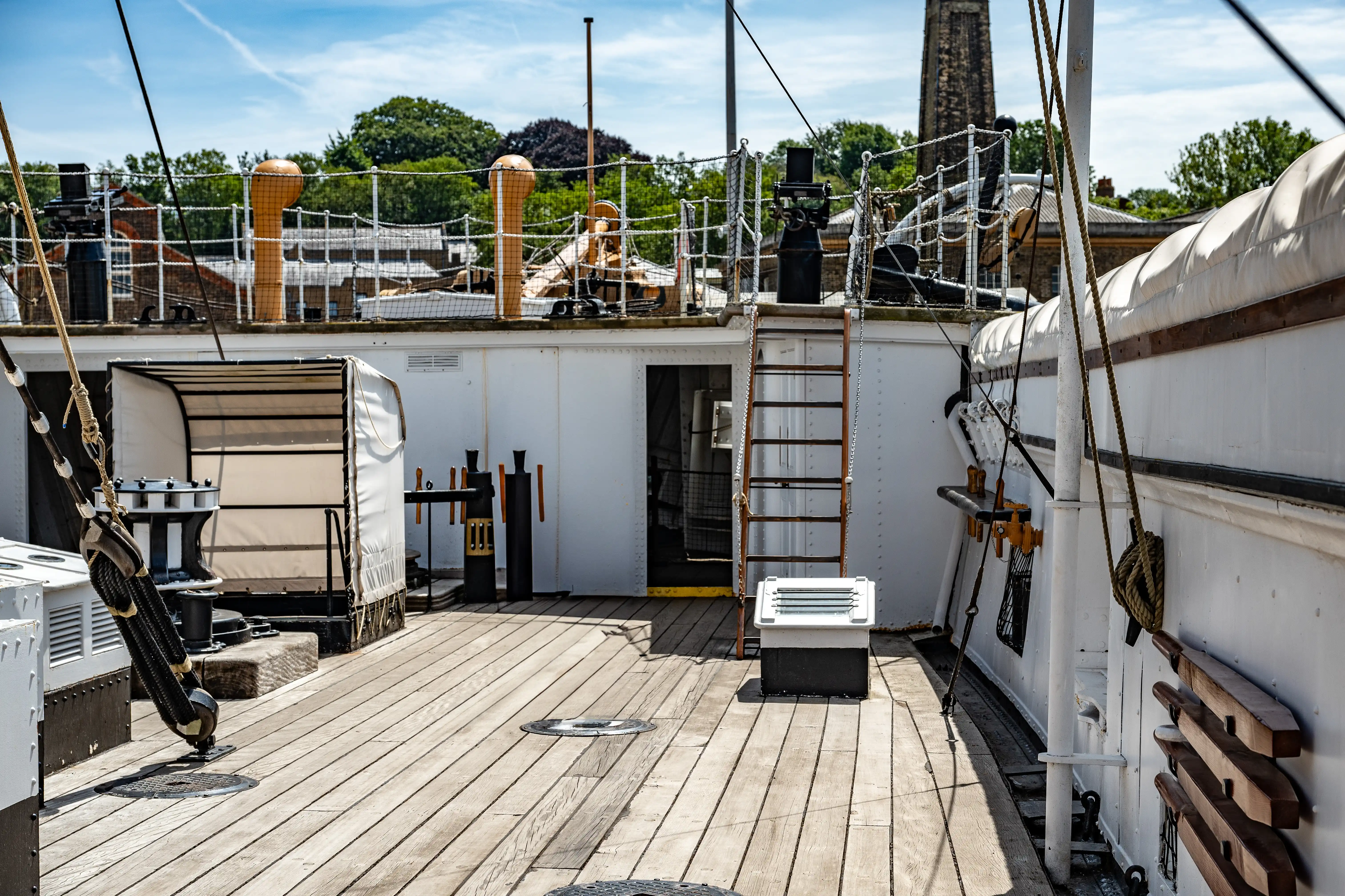 The image depicts a section of a ship's deck. The deck is made of wooden planks and features various maritime equipment and structures, including covered lifeboats, ventilation shafts, and ropes. There is a ladder leading to a higher deck level, and the ship's railings are visible. In the background, there are industrial buildings and greenery, indicating the ship might be docked in an urban area.