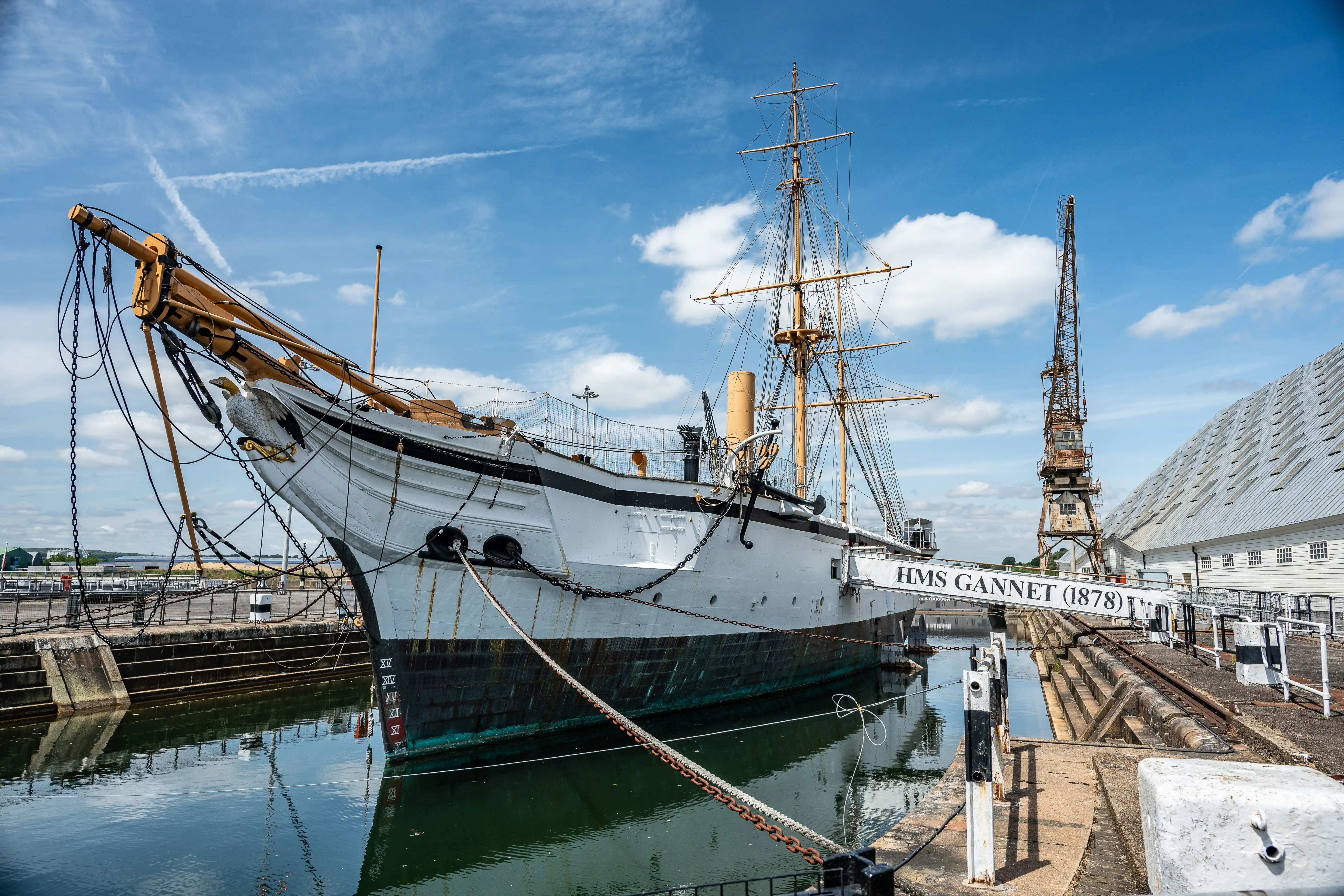 The image depicts a historic ship named HMS Gannet, built in 1878, docked in a harbor. The ship is moored with chains and ropes, and a figurehead of a bird is visible at the bow. The background includes a crane and a modern building, indicating a blend of historical and contemporary elements in the setting. The sky is clear with some clouds, suggesting a bright day.