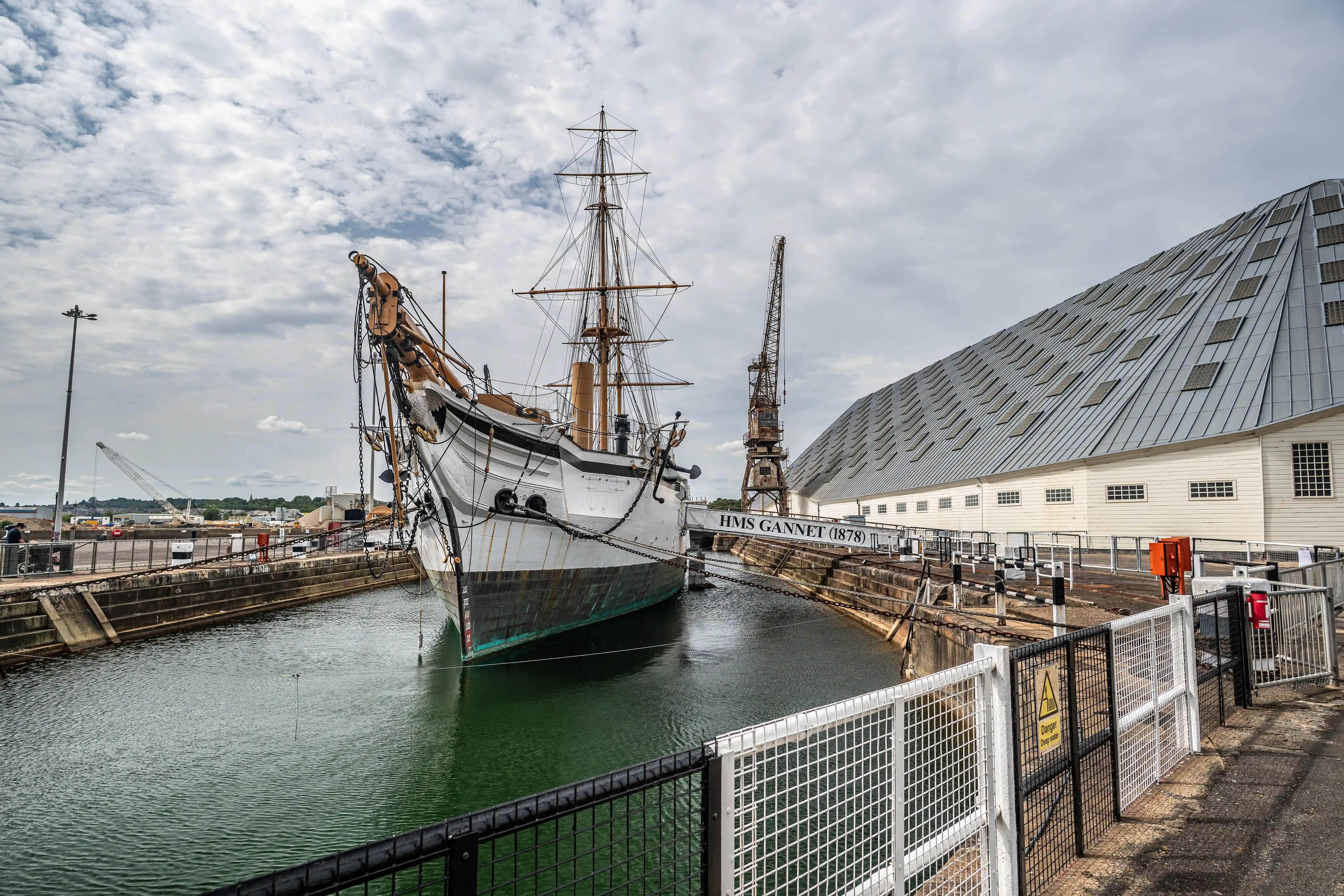 The image depicts a historic ship named HMS Gannet (1878) docked in a dry dock. The ship is a well-preserved example of maritime history, with visible masts and rigging. The surrounding area includes modern infrastructure such as a large metal building and a crane, indicating a blend of historical preservation and contemporary development. The scene is set under a cloudy sky, adding a dramatic backdrop to the historical vessel.