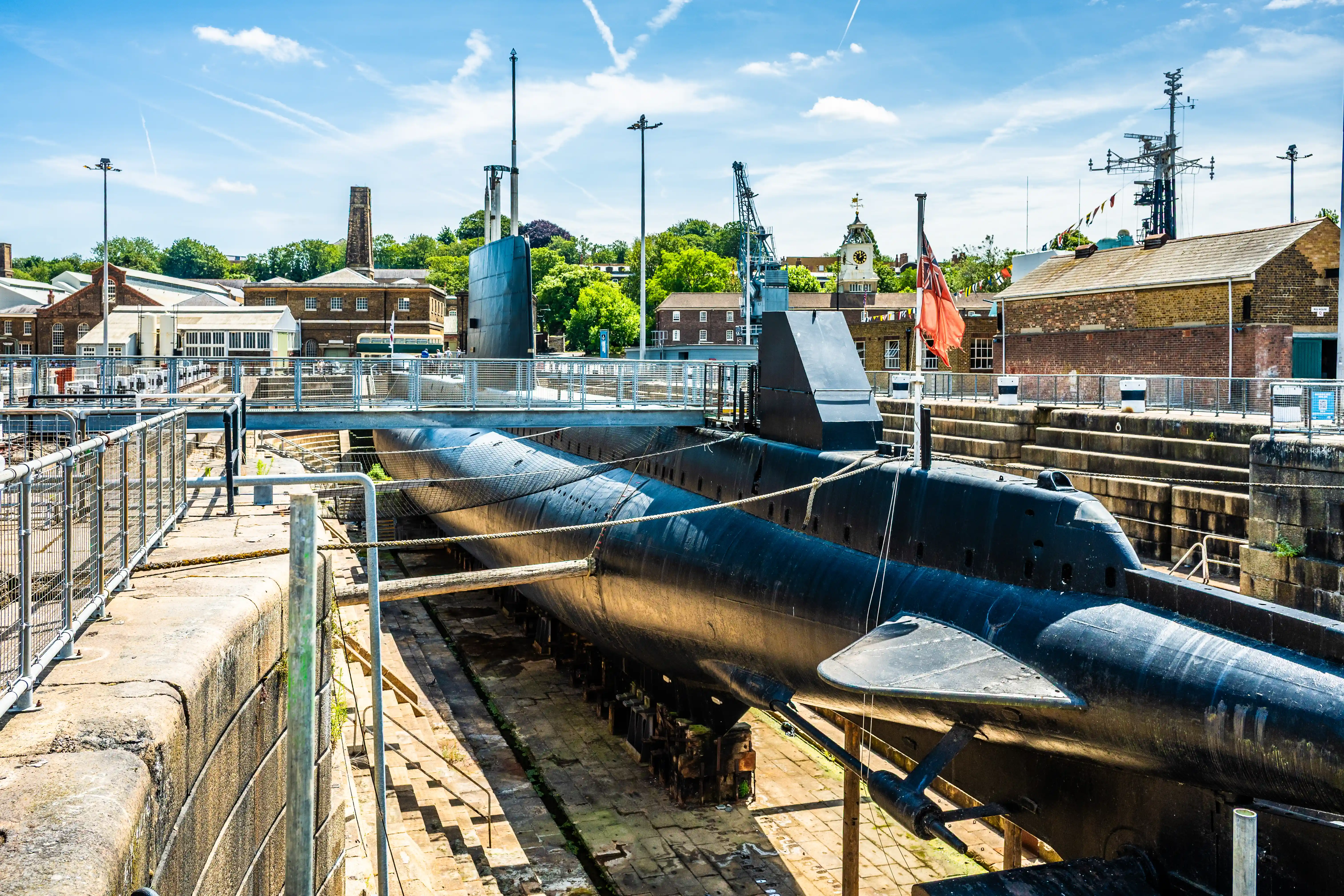 The image depicts a dry dock area with a submarine in the foreground. The submarine is elevated and supported by wooden blocks, indicating it is undergoing maintenance or is on display. Surrounding the dock are various buildings, likely part of a historical or maritime museum complex. A flag is visible near the submarine, and there are railings and pathways for visitors. The background shows additional maritime structures and greenery under a clear sky.