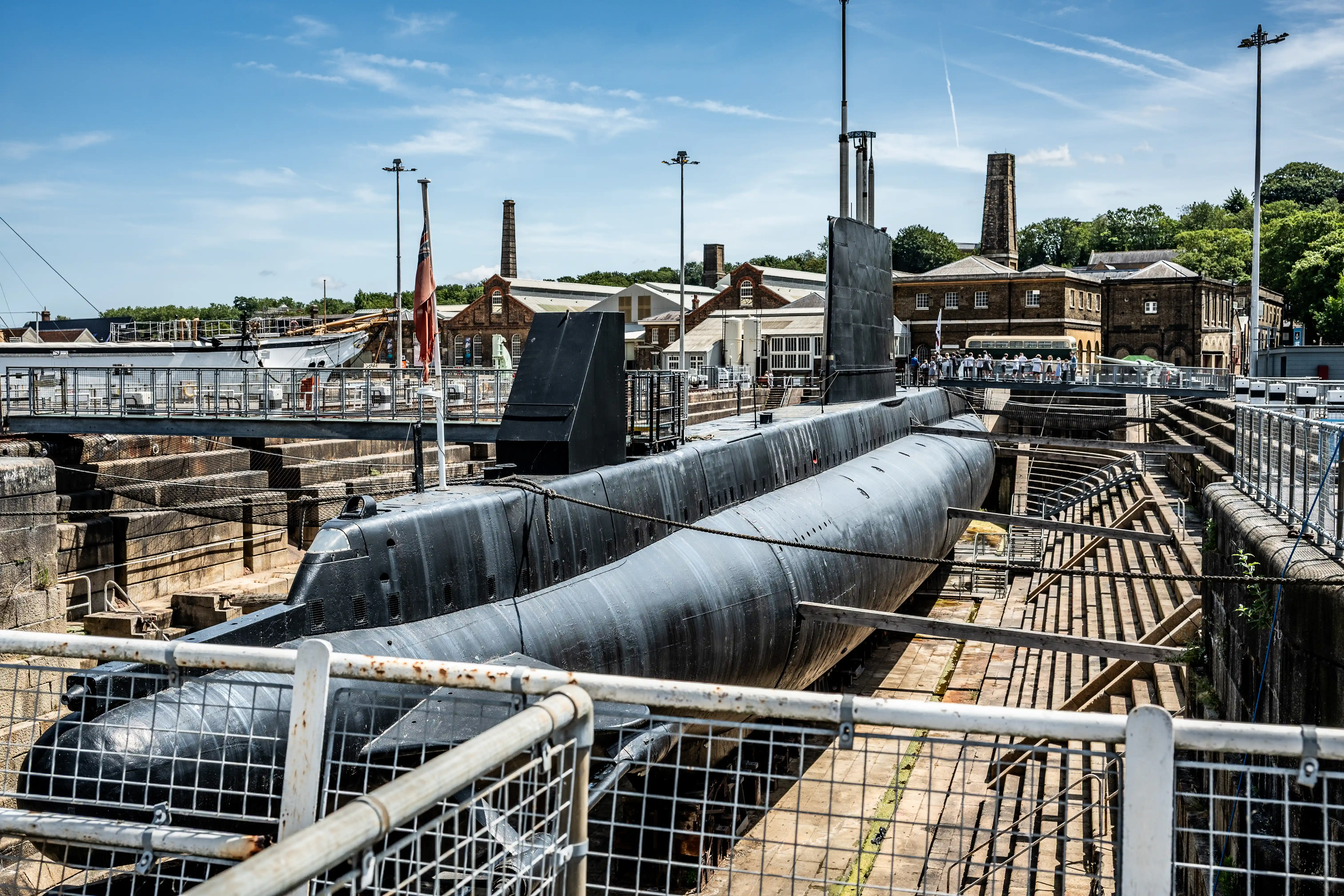 The image depicts a large submarine docked in a historic shipyard. The submarine is situated in a dry dock, surrounded by industrial buildings and structures. There are walkways and railings around the dock area, with a few people visible in the background. The scene is set on a clear, sunny day, highlighting the details of the submarine and its surroundings.