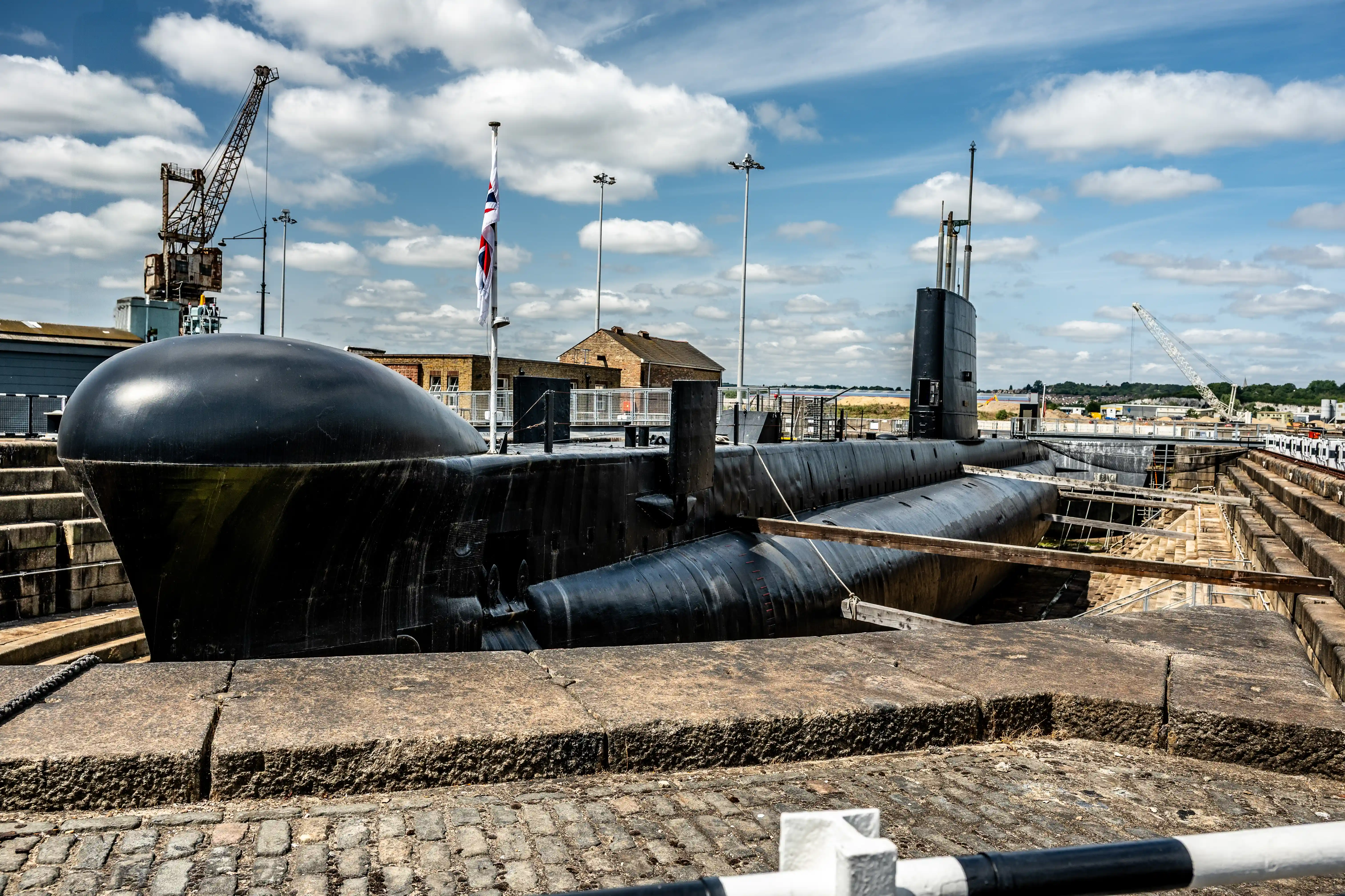 The image depicts a submarine docked in a dry dock. The submarine is black and appears to be in a stationary position. The surrounding area includes industrial structures such as cranes and buildings, with a partly cloudy sky overhead. The scene suggests a naval or shipyard setting.