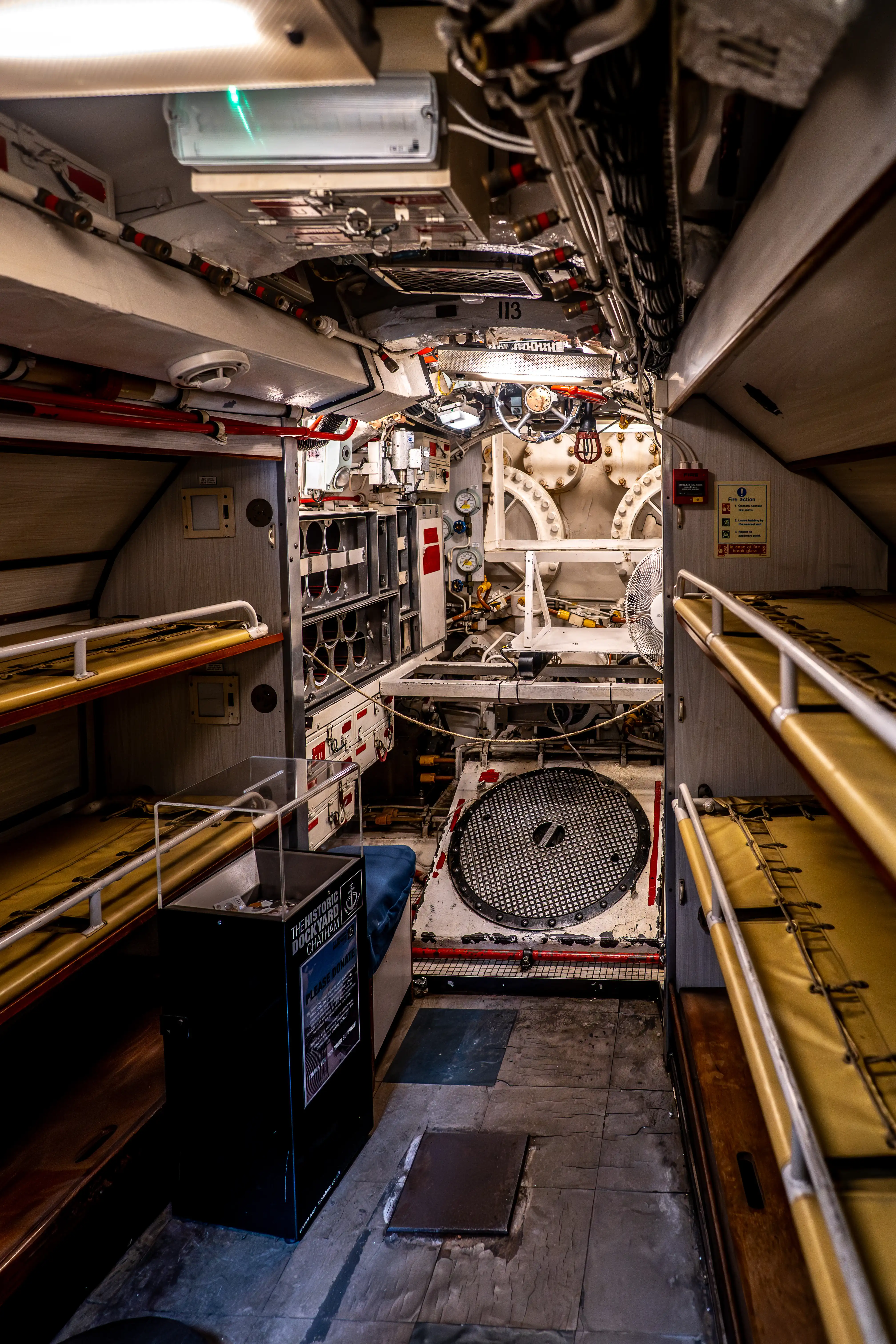 The image depicts the interior of a submarine, showcasing a narrow corridor with various equipment and machinery. The area is compact with multiple pipes, gauges, and control panels visible. There are bunk beds on either side of the corridor, indicating sleeping quarters for the crew. Safety and operational instructions are posted on the walls, and the floor appears to be made of metal grating. The environment suggests a highly functional and industrial setting designed for underwater operations.