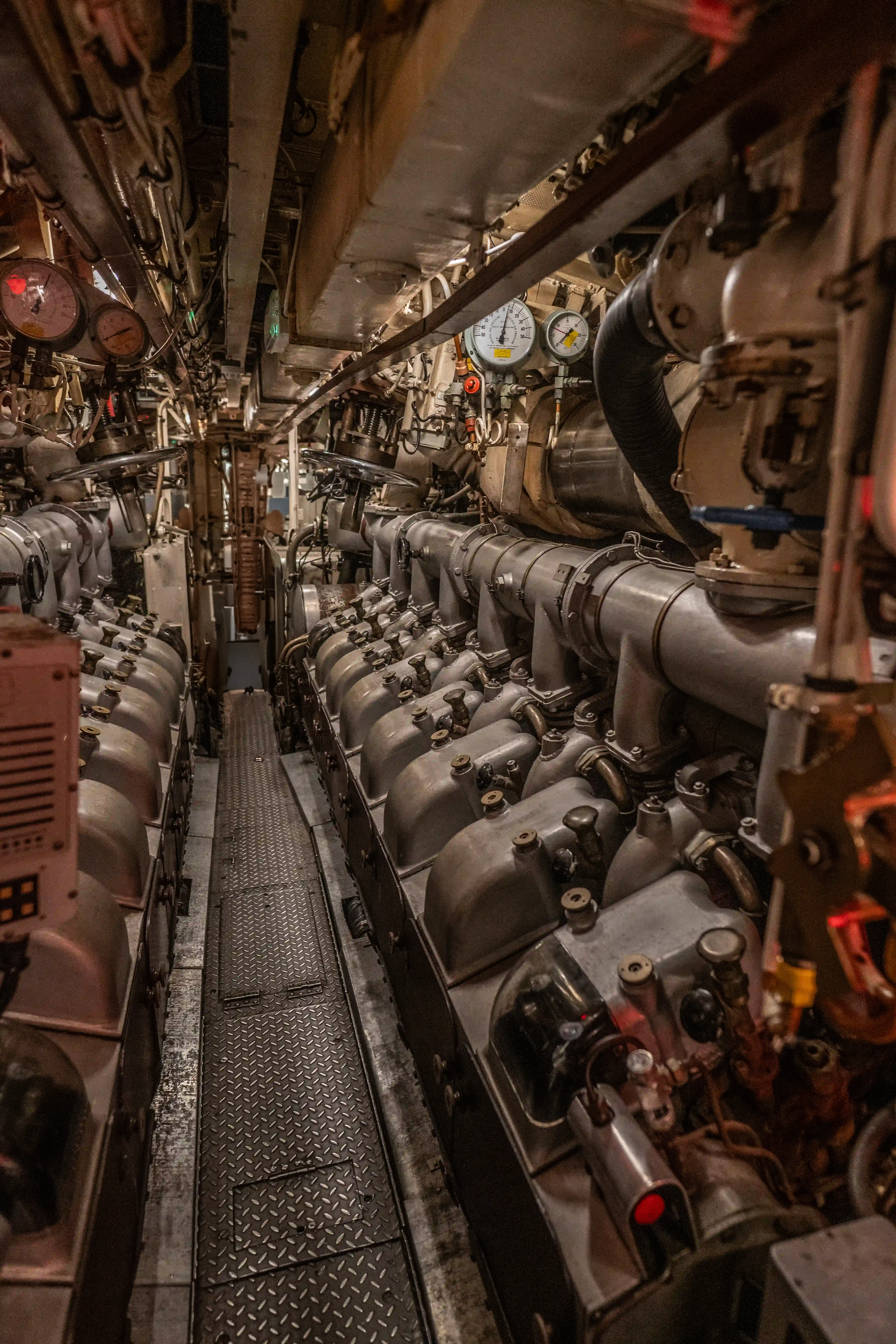 The image depicts the interior of an engine room, likely from a ship or submarine. It features a complex array of machinery, including multiple engines, pipes, gauges, and various mechanical components. The environment appears industrial and tightly packed with equipment, indicative of a space designed for functionality and efficiency. The flooring consists of metal grating, and the overall scene suggests a well-maintained but heavily used mechanical area.